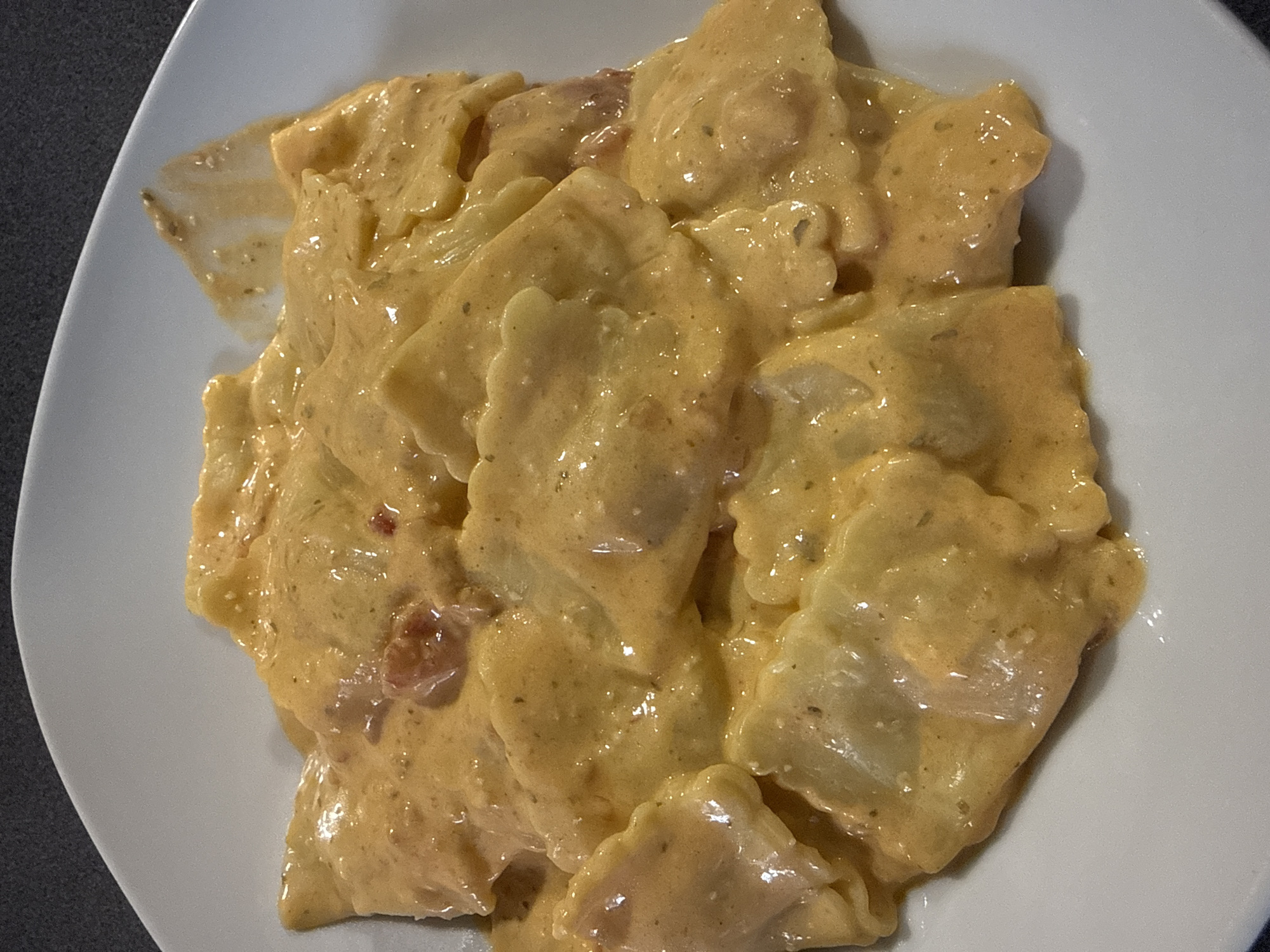 A close-up, overhead shot displays a white, shallow bowl filled with a generous portion of ravioli. The pasta is coated in a creamy, orange-hued sauce, likely a tomato-based cream sauce, with visible flecks of herbs and possibly small pieces of tomato. The ravioli themselves are square-shaped with scalloped edges, and they are arranged in a layered fashion, partially overlapping each other. The lighting highlights the sheen of the sauce, giving the dish a rich and appetizing appearance. The background is a dark, textured surface, likely a countertop or table, which contrasts with the bright white bowl and the warm colors of the food. There are no people or any discernible urban elements present in the image, and therefore no information about the city of Marum, Netherlands can be inferred.