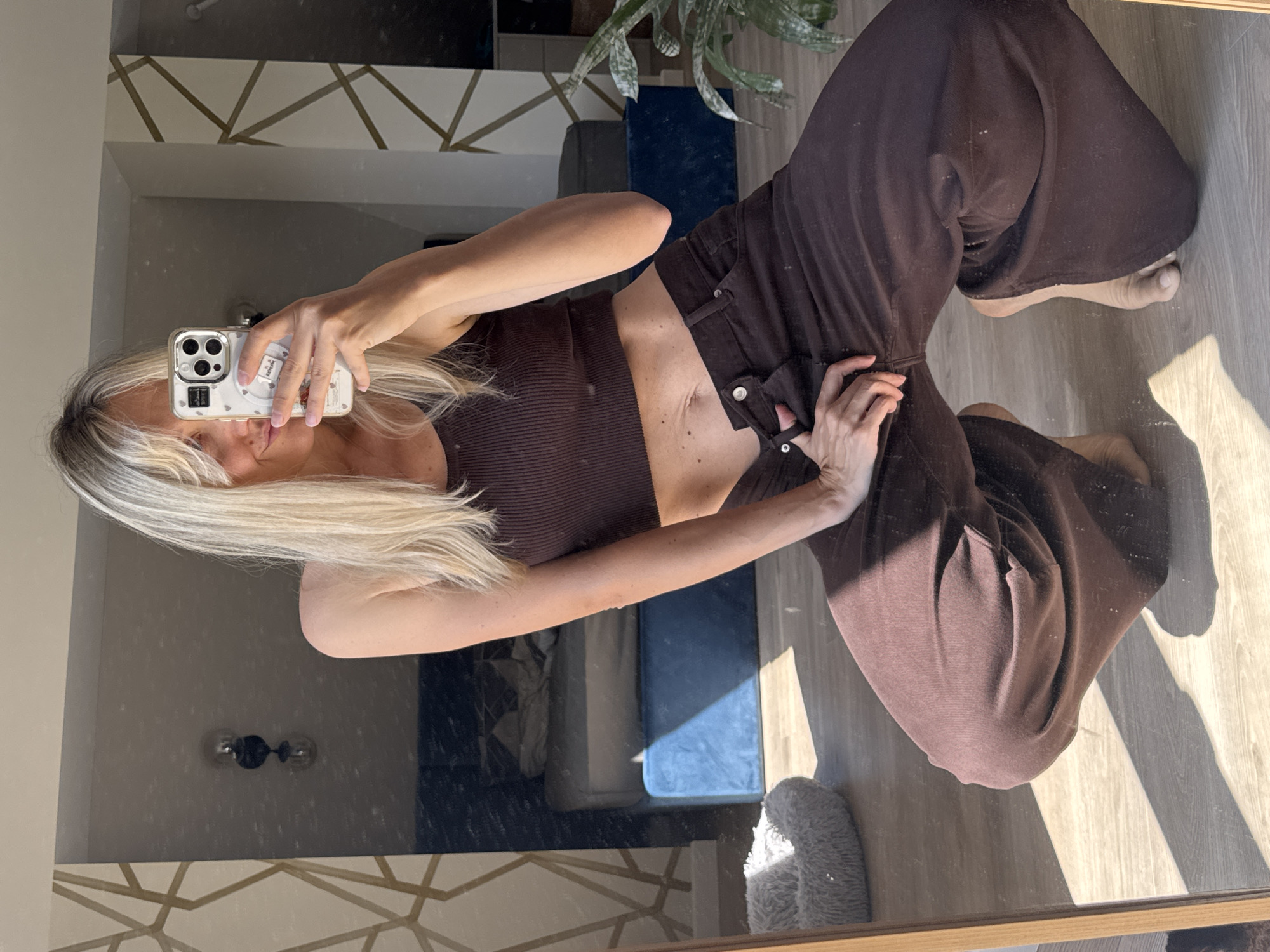 A woman with blonde hair is crouching in a room, holding a smartphone to take a selfie in a mirror. She is wearing a brown ribbed tank top and brown wide-leg pants. The room appears to be a bedroom with a bed in the background. Sunlight streams in from a window, casting shadows on the wooden floor. There is a dog bed on the floor to the left, and a plant to the right. The background shows a wall with a geometric pattern and a lamp. The image quality is good, with clear focus and natural lighting. No specific location in Carnikava, Latvia is identifiable.