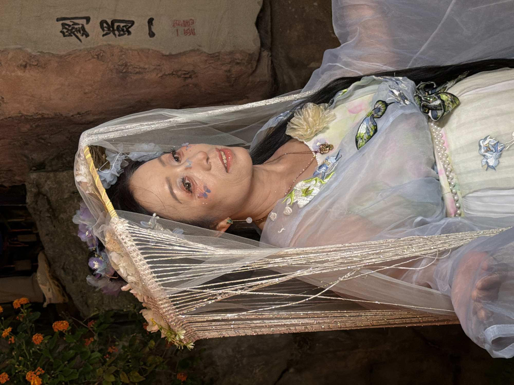 In a dimly lit, outdoor setting possibly in Yuzhong District, China, a middle-aged East Asian woman is captured in a medium shot. She is adorned in an elaborate, ethereal costume featuring sheer, pale blue and white fabric with embroidered floral and butterfly motifs. A delicate, shimmering veil with dangling beaded strands covers her head and shoulders, adorned with flowers and pearls. Her makeup is dramatic, with colorful accents and glitter applied to her eyelids and cheeks. She wears a necklace with a pendant. The background consists of textured rock formations and foliage with orange flowers. To the right of the woman, a stone wall bears vertical Chinese calligraphy. The lighting is dim, with some light highlighting the woman and her attire.
