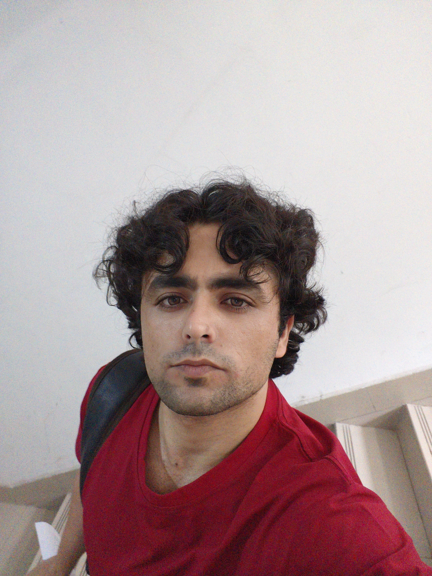 A medium shot selfie features a man from the chest up. He has dark, curly hair and brown eyes. The man is wearing a red t-shirt and a dark backpack strap is visible over his left shoulder. The background is a plain white wall and a staircase. The image appears to be taken indoors.