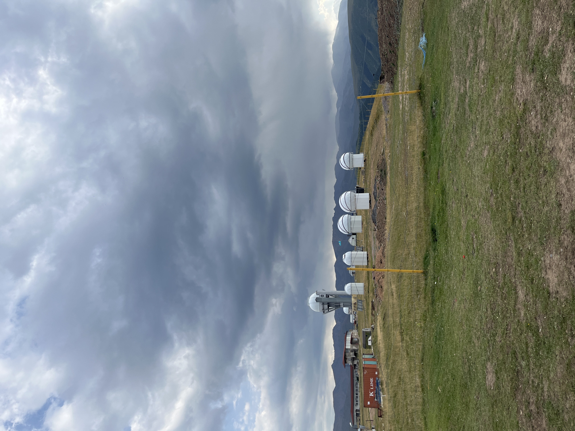 The image shows an observatory with several white dome-shaped structures and a tall tower with a dome on top, situated on a grassy hillside. In the background, there are mountains and a cloudy sky. There are also some buildings and shipping containers to the left of the observatory domes. The scene appears to be an outdoor, mountainous landscape, possibly an astronomical research facility. The location in Batan, Kazakhstan, suggests a remote, high-altitude setting suitable for astronomical observation.