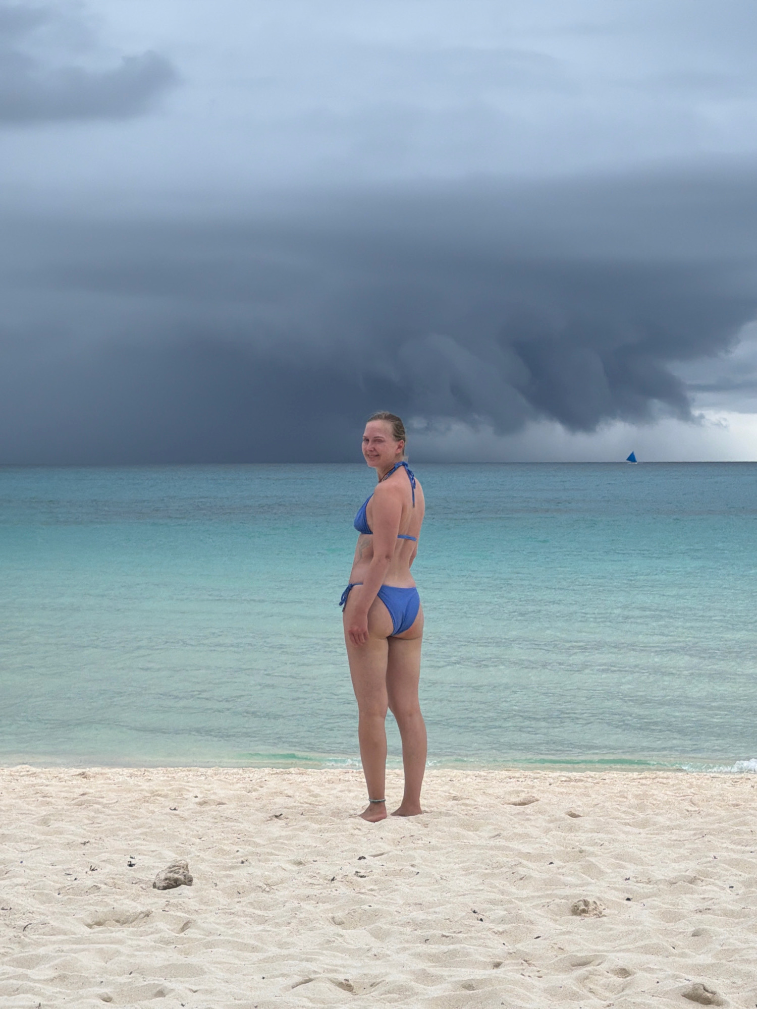 A woman in a blue bikini stands on a sandy beach, facing the ocean and looking over her shoulder at the camera. The ocean is a clear turquoise color, and in the distance, a dark storm cloud looms over the horizon. A small sailboat is visible on the water near the horizon. The beach is covered in white sand with some small debris. The scene appears to be the city of Malay, Philippines, possibly Boracay Island, given the white sand and turquoise waters.