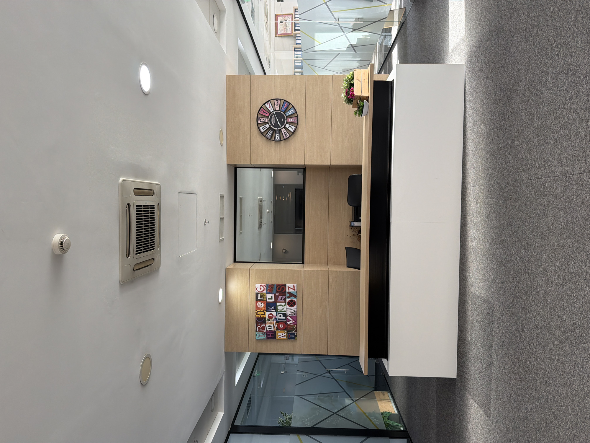 The image depicts the interior of a modern building, possibly an office or commercial space, located in Sofia, Bulgaria. A light-colored wall is visible on the left, featuring a circular recessed light, a square air conditioning vent, a smoke detector, and a small rectangular access panel. A wooden structure occupies the central portion of the image, incorporating a clock, a square panel with alphabet letters, a glass partition, and a small green potted plant. To the right of the wooden structure, there is a white, rectangular structure. The floor is carpeted in a medium gray tone. The ceiling has reflective glass panels and geometric beams. There are no people visible in the image.