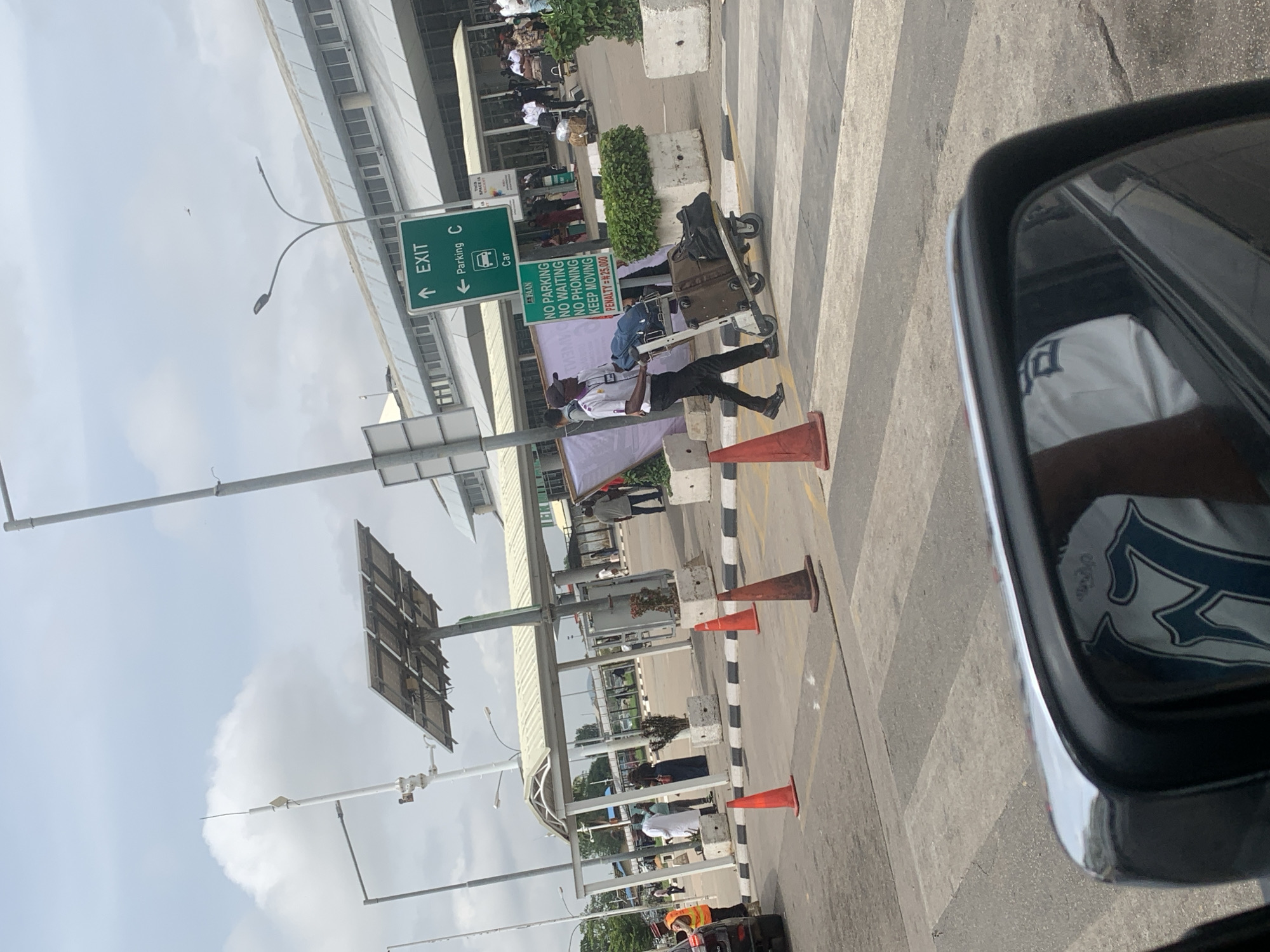 The image, taken from inside a car, depicts a busy outdoor area at what appears to be an airport in Gwaska, Nigeria. The scene is set during the daytime, under a bright sky with scattered clouds.
In the foreground, a car's side mirror is visible on the right, reflecting part of the car's interior. A pedestrian crosswalk with alternating light and dark grey stripes stretches across the asphalt road. Several orange traffic cones are placed along the edge of the road, delineating a path or restricted area.
The central subject is a man, dressed in a light-colored collared shirt and dark trousers, actively pushing a metal luggage cart laden with bags. He walks along the crosswalk, seemingly moving towards or away from a building. Behind him and further in the background, various people are visible, some appearing to wait or walk near a building facade, which suggests an airport terminal. Concrete planters with greenery and large concrete blocks are also present.
Overhead, multiple street lights and poles with mounted equipment, possibly solar panels, are visible against the sky. A prominent green road sign indicates "↑ EXIT" and "← Parking C" with a "Car" icon. Below it, another green sign displays regulations: "FAAN", "NO PARKING", "NO WAITING", "NO PHONING", "KEEP MOVING", and warns of a "PENALTY = #25,000", confirming the Nigerian context. The overall activity suggests a dynamic environment typical of an airport drop-off or pick-up zone.
