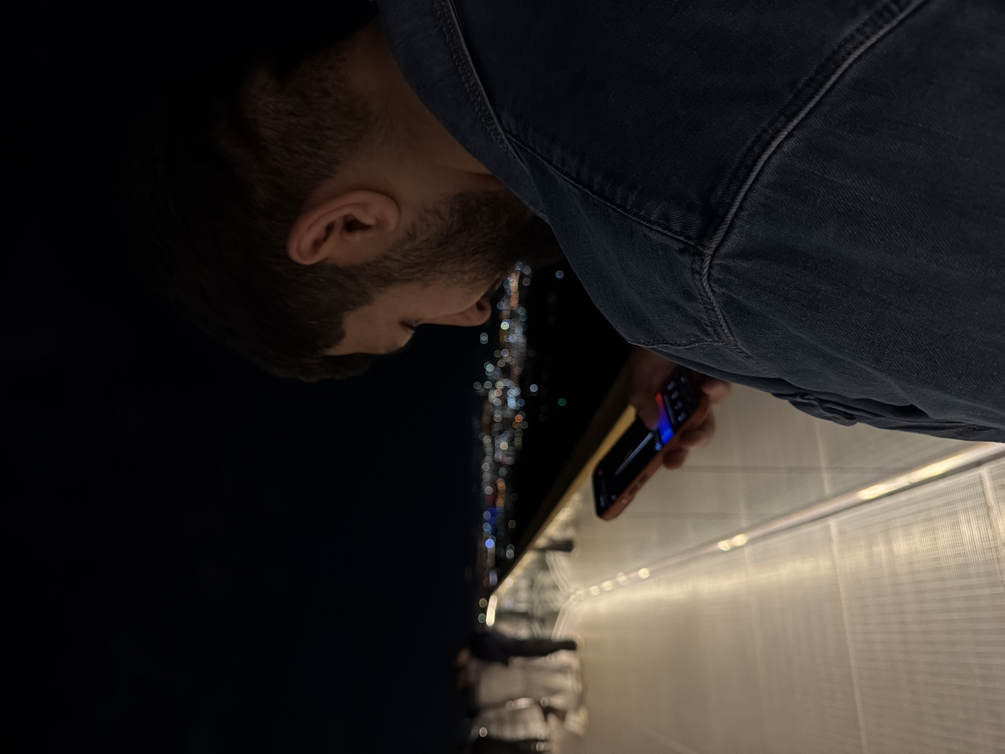 A man in a dark blue denim jacket is standing on a brightly lit walkway, possibly made of translucent material, at night. He is holding a smartphone in his right hand, looking down at it. In the background, there is a blurry, bokeh-style view of city lights, indicative of Krasnoyarsk, Russia. The perspective suggests the man might be elevated, possibly on a bridge or high walkway overlooking the city.