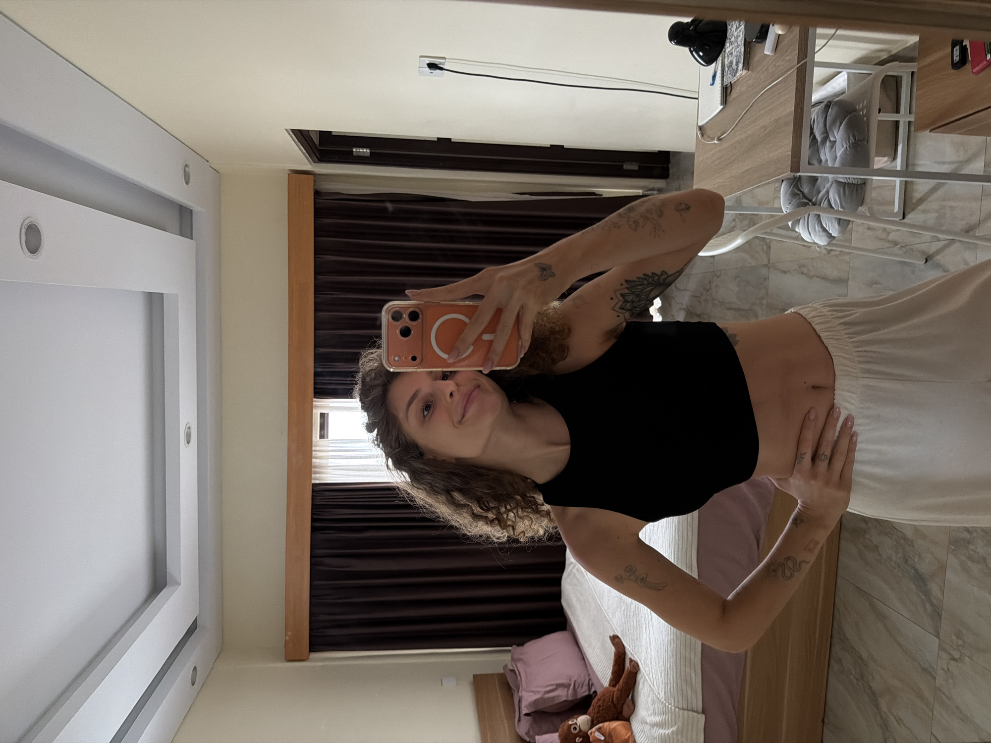 A young woman with curly brown hair is taking a mirror selfie in what appears to be a bedroom in North Kuta, Indonesia. She is wearing a black crop top and loose white pants. She has several tattoos on her arms, including a flower mandala and a snake. She is holding an orange smartphone in her right hand.
The room has a bed with a pink headboard and two pillows, one of which has a stuffed monkey toy on it. There are dark brown curtains with a lighter fabric behind them, and a white-framed window. The walls are a light beige color, and the ceiling has a recessed lighting design. To the right of the woman is a desk with a lamp, a notebook, and other items. The floor is tiled with a marble pattern. The overall impression is of a brightly lit, casual indoor space.