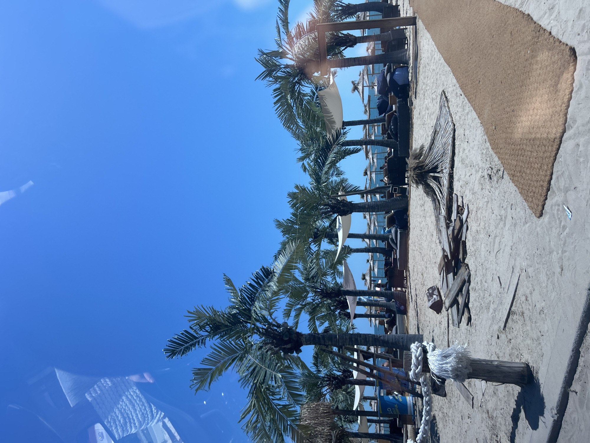 This is a tilted, full shot of a beachside area on a sunny day, likely in Varna, Bulgaria. The scene is dominated by palm trees, some appearing artificial, lining a sandy area. A rough, tan-colored mat is laid out on the sand, diagonally across the frame. In the background, there's a structure resembling a beach bar or lounge area with wooden beams and white coverings, and several people are seated or standing within this area, though their features are not clearly discernible. A wooden post with a thick white rope tied around it stands prominently in the foreground on the sand. Scattered debris, including pieces of wood, are visible on the sand. The sky is a clear, bright blue, indicating good weather and likely midday. The overall mood is relaxed and tropical, suggesting a vacation or leisure setting. There is no visible text in the image.