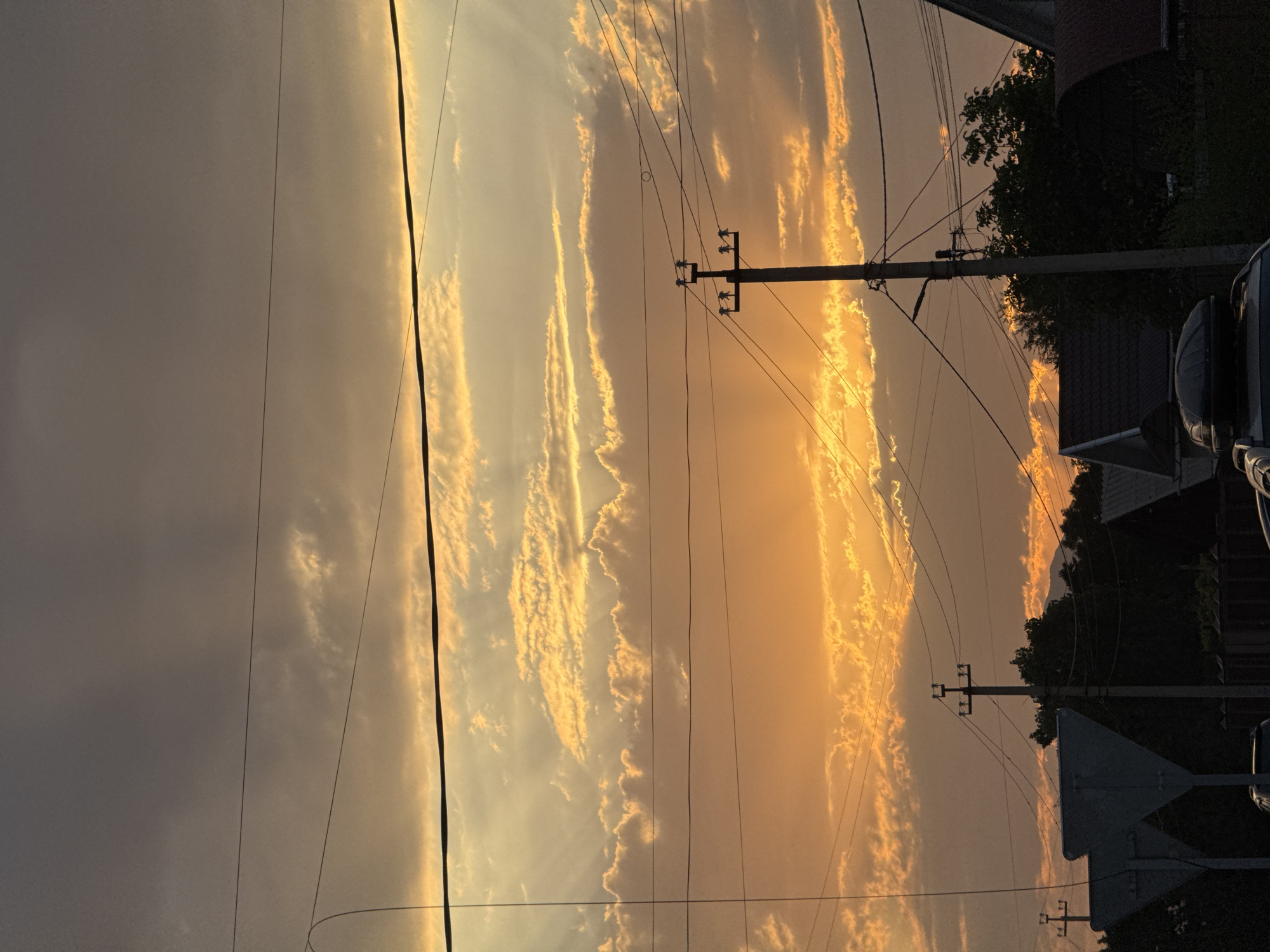 A full shot captures a dramatic sunset over a suburban area of Talas City, Kyrgyzstan. The sky is dominated by vibrant orange and yellow hues, with layered clouds reflecting the warm light. Rays of sunlight pierce through gaps in the clouds, creating a striking visual effect. Overhead power lines crisscross the frame, interrupting the sky. In the foreground, silhouettes of houses, trees, and a utility pole are visible against the brightly lit sky. The scene appears to be a residential street with some traffic signs and a car roof box in the lower portion of the image. The overall atmosphere is serene and highlights the natural beauty of the sky during sunset.