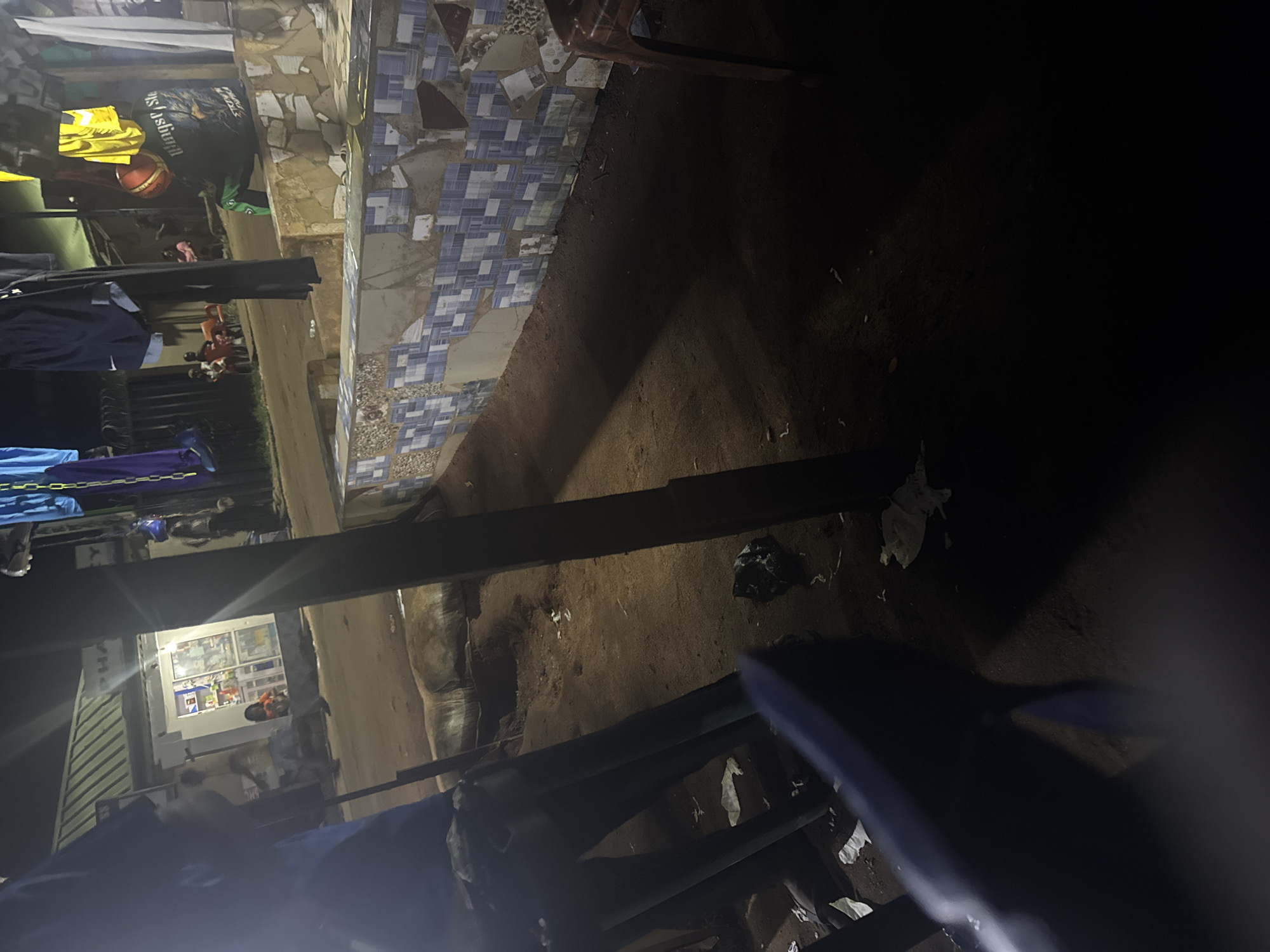 The image depicts a nighttime scene in Ihiagwa, Nigeria, focusing on a street-level view. In the foreground, a dark blue chair is partially visible, its form distorted by the low light. To the left, a person in dark clothing stands behind what appears to be a collection of chair legs. The ground is a mix of dirt and scattered white debris, suggesting a somewhat unkempt area.
Further back, a small shop or establishment with a green corrugated roof is illuminated by interior lights, revealing shelves stocked with various items, possibly pharmaceuticals or convenience goods. Several individuals are present, including a child in an orange shirt and another person in a yellow shirt and blue shorts. They are standing near the shop entrance.
To the right, a person wearing a dark jacket with lettering that reads "Wings of Steel" and green pants is seated on a low, tiled wall. They have a red object, possibly a basketball, resting on their lap. Above them, laundry is hung on a line, including blue and yellow garments. A wooden stool or chair is positioned to the far right. The overall environment appears to be a community gathering or commercial area at night, with artificial lighting illuminating the immediate surroundings.