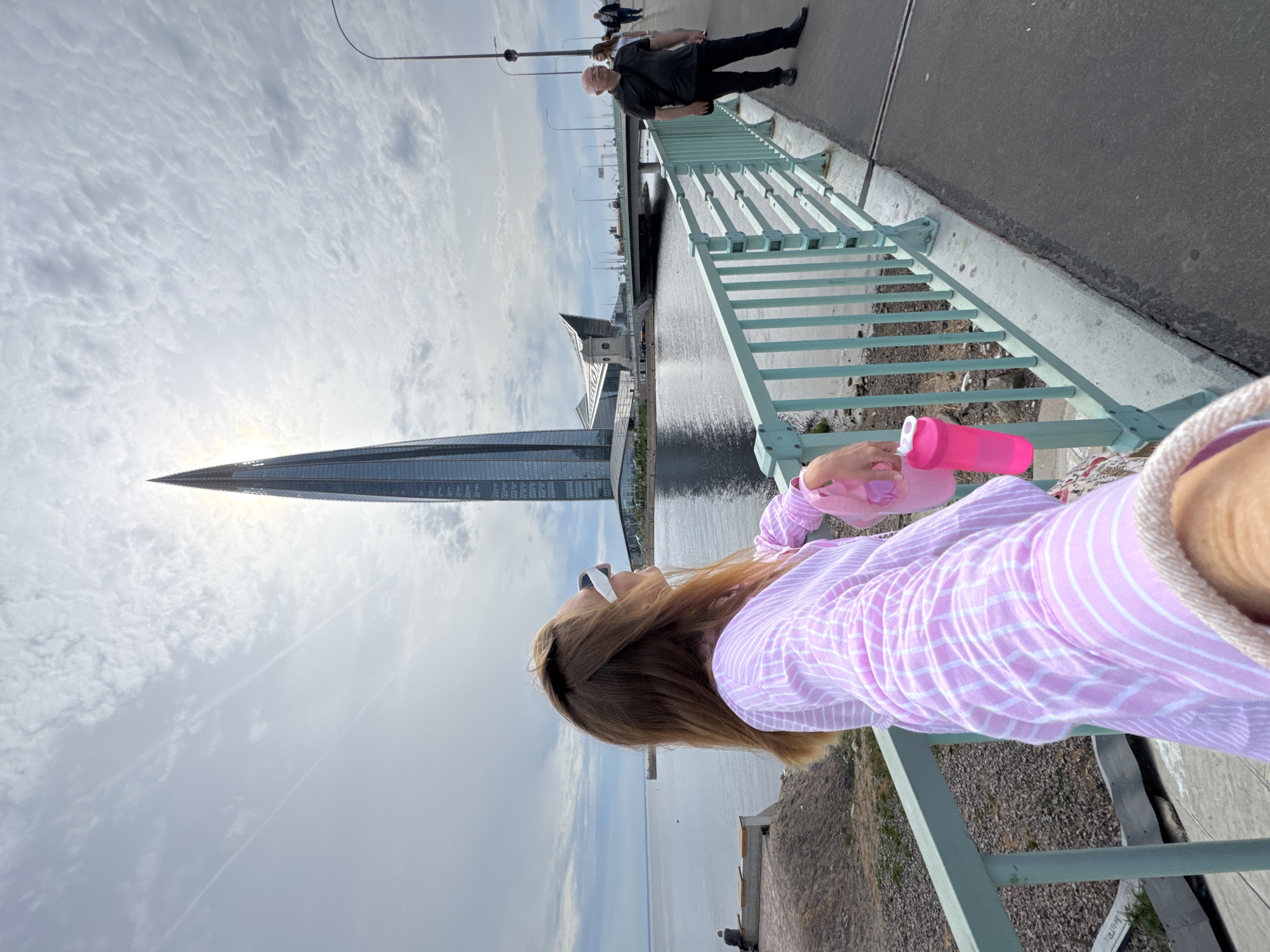 The scene is a bridge in Saint Petersburg, Russia, with a tall skyscraper, the Lakhta Center, visible in the background. The photograph, taken from an elevated perspective, depicts a person holding a pink water bottle or container, positioned near the bridge's railing. Other people are walking on the bridge in the background. The bridge railing is light green and the water below the bridge is dark. The sky is overcast with clouds.