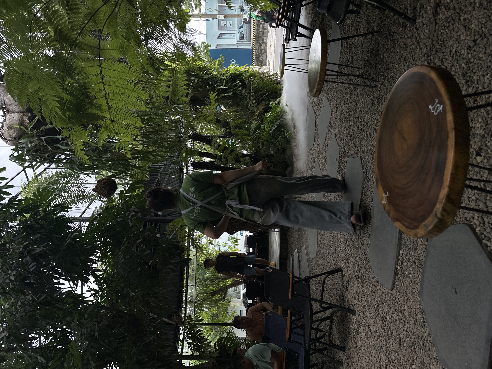 The image captures an outdoor seating area of a cafe in Da Nang, Vietnam, lush with tropical plants and ferns. A person in a green apron and light-colored pants, seemingly an employee, is walking away from the camera, with a subtle mist rising from the ground around them. Several empty tables and chairs are scattered across a gravel ground, interspersed with flat stone pavers. In the background, a man in a dark sleeveless shirt and shorts walks on a wooden platform, and other patrons are seated at tables. The cafe's environment is verdant and open-air, suggesting a relaxed, nature-inspired atmosphere.