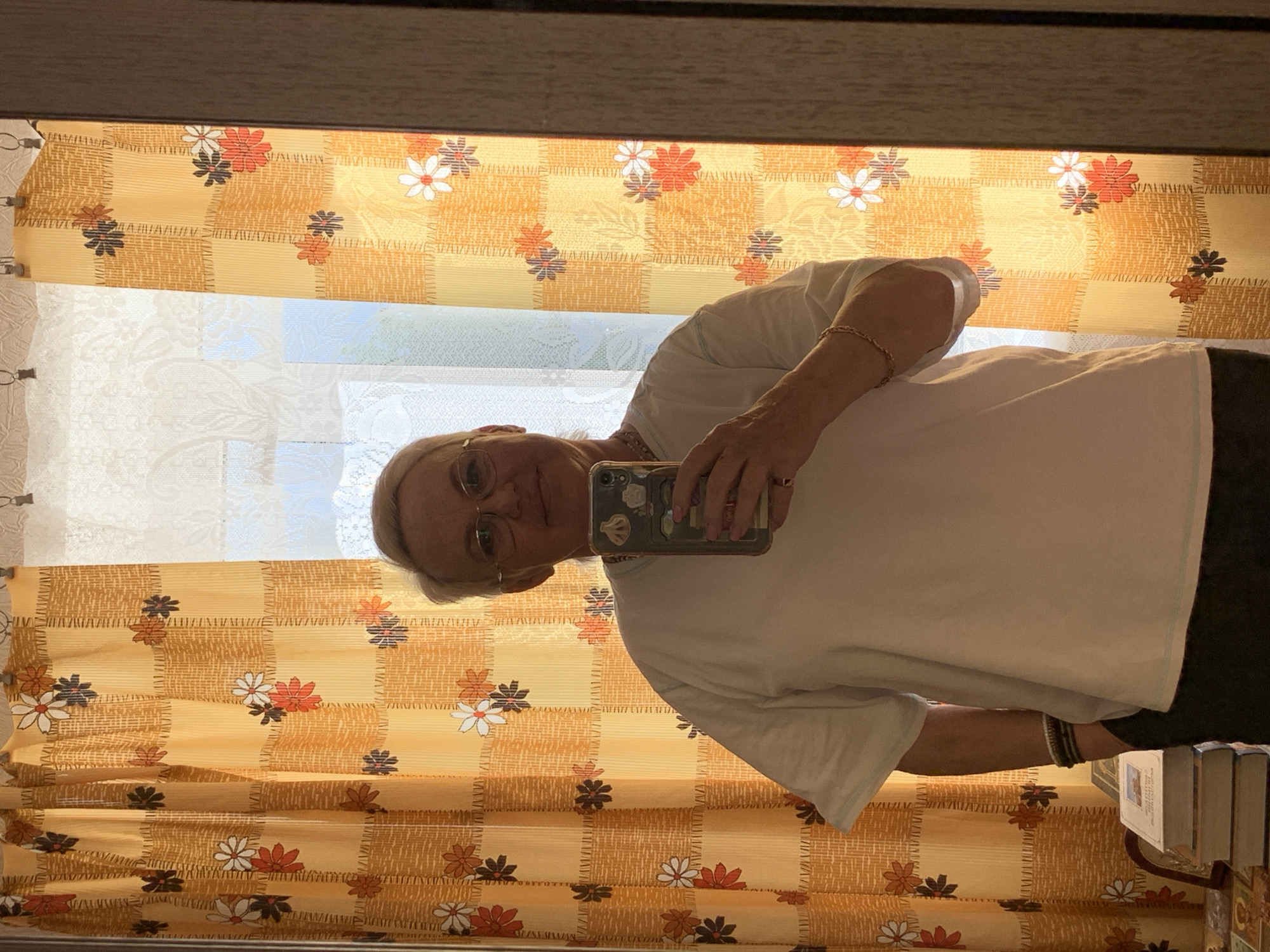 A woman in a white T-shirt is taking a mirror selfie with her smartphone. She is wearing glasses and jewelry on her wrists. The background includes a window with a patterned curtain and a stack of books to the right. The location is inferred to be in Chaikovsky, Russia.