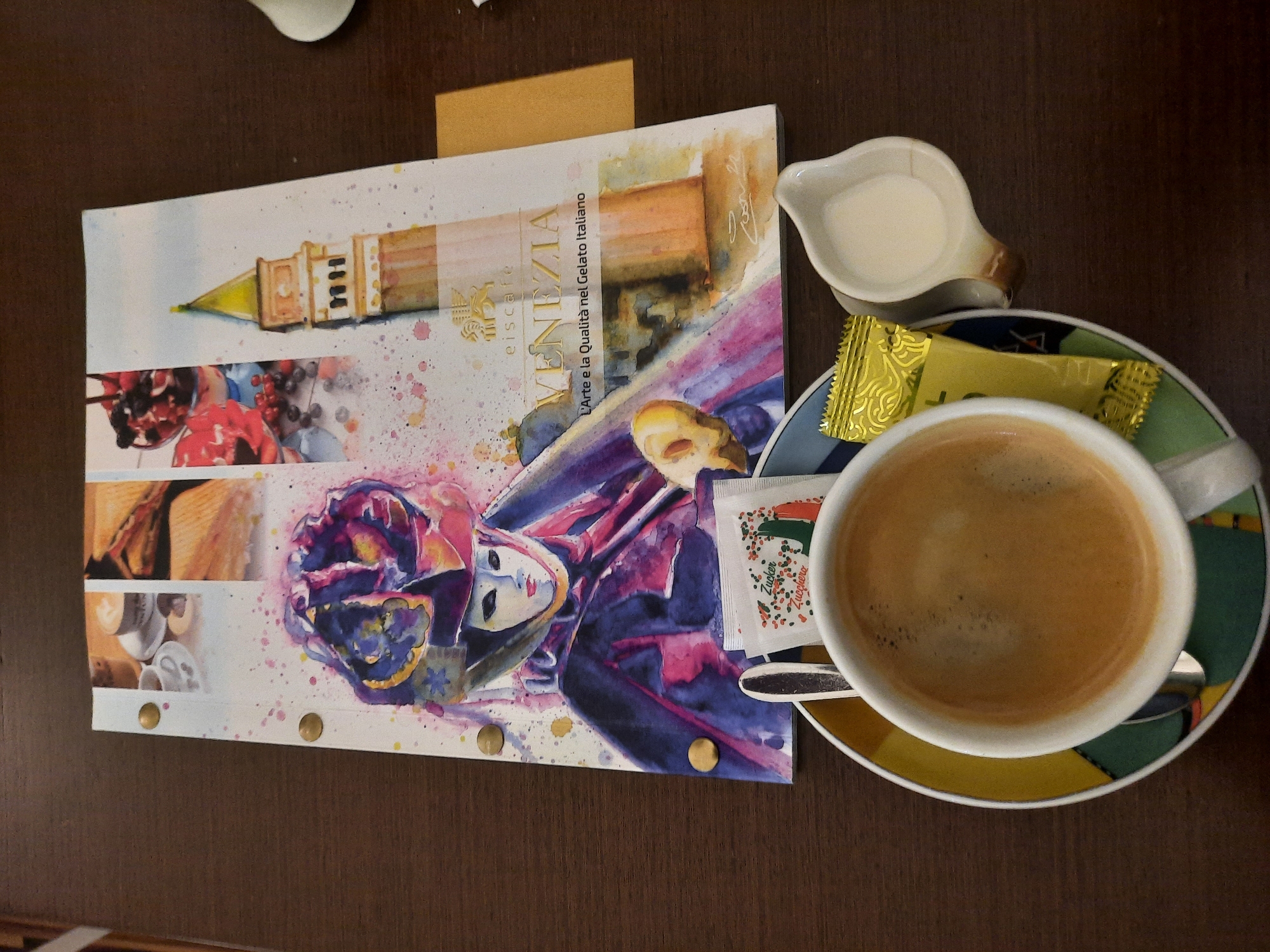 A top-down medium shot captures a table with a coffee cup, creamer, sugar packet, and a menu. The coffee is in a white mug with a crema layer and sits on a saucer with a geometric pattern in yellow, blue, and green. Next to the cup is a small white ceramic creamer filled with milk and a gold foil sugar packet. A menu, featuring watercolor artwork of a Venetian masquerade figure and a bell tower, lies open to the right of the coffee. The menu has the text "eiscafé VENEZIA" and "L'Arte e la Qualità nel Gelato Italiano" printed in gold. The table surface is a dark wood grain. The setting is indoors, likely a cafe, given the menu and coffee service. No people are visible in the image. The image contains no identifiable landmarks or features that would specifically place it in Rosenheim, Germany.