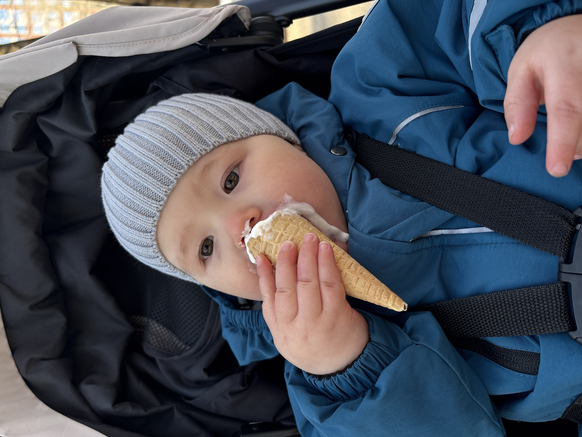 A young child wearing a grey knitted hat and a blue winter jacket sits in a stroller, eating an ice cream cone. The child's hand holds the cone, with ice cream smeared around their mouth. The stroller has a light-colored canopy and a black interior. Black straps are visible across the child's jacket. The scene appears to be outdoors. There is no evidence to suggest that the location is Izhevsk, Russia.