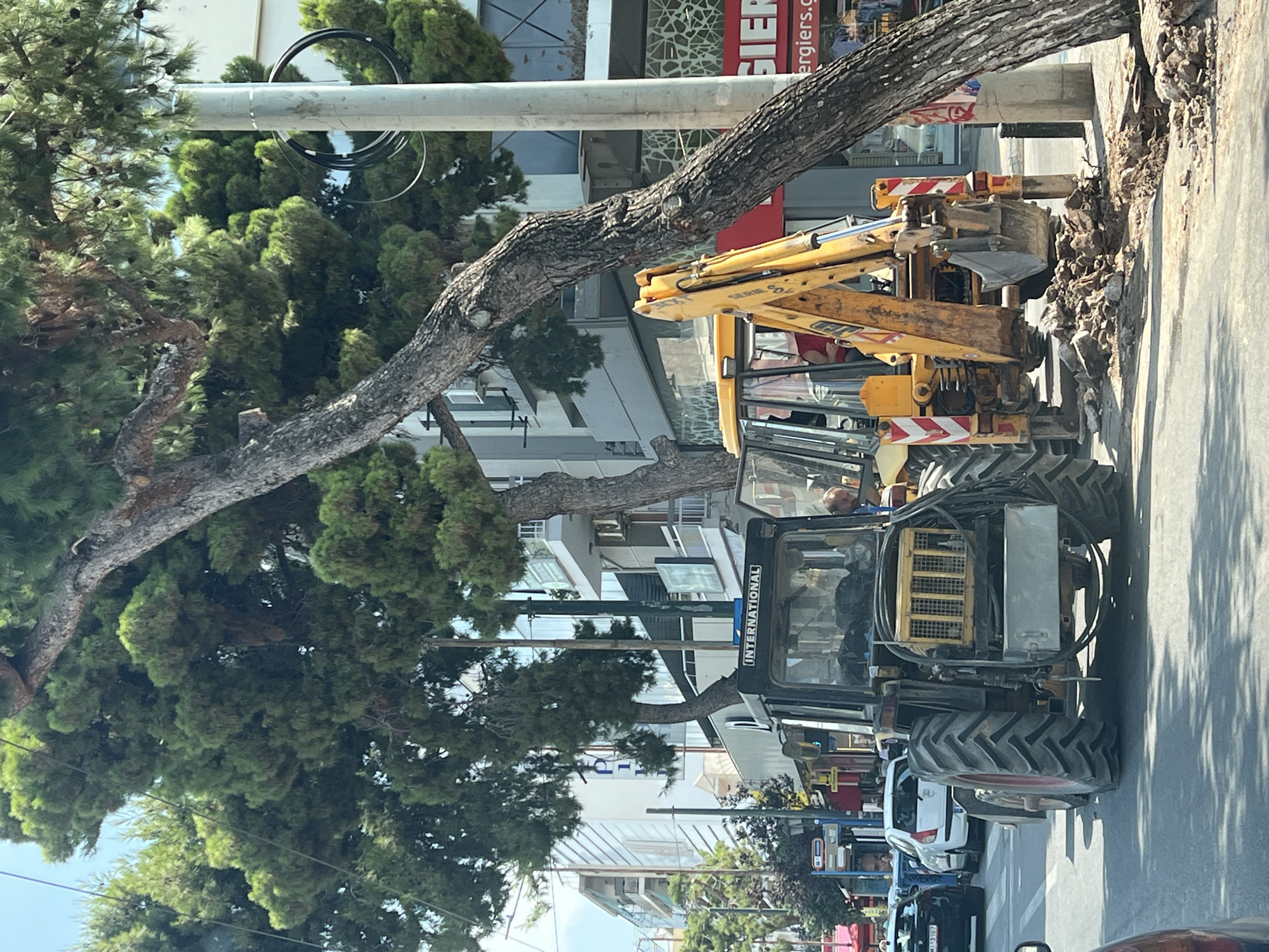 In Kallithea, Greece, on a sunny day, a street excavation is in progress. Two construction vehicles are positioned on the paved road. In the foreground, a black tractor with the "INTERNATIONAL" logo is visible. Behind it, a yellow backhoe excavator is partially obscuring it. The excavator's bucket is lowered into a freshly dug pit on the side of the road, with piles of dirt and rocks around it. A large pine tree with a thick trunk extends from the right side of the frame, casting shadows over the scene. The background reveals urban elements: multi-story buildings with balconies, shops, streetlights, and other vehicles, including a white car and a black car, suggesting a bustling city environment. The sky is clear and blue.