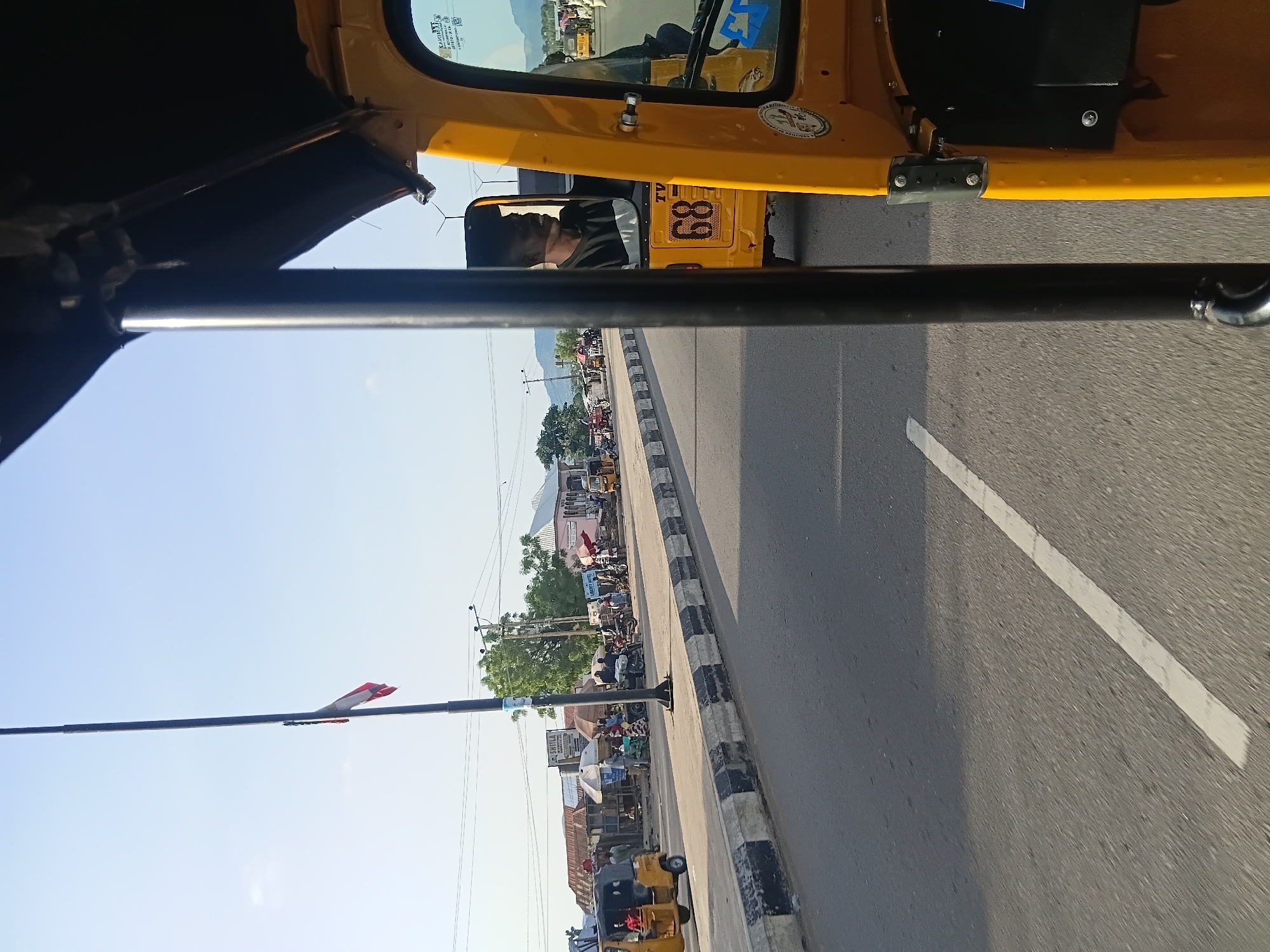 In Jalingo, Nigeria, a view from inside a yellow auto-rickshaw traveling on a paved road shows a street scene. The auto-rickshaw, with a black metal roll cage and a black canopy, is in the foreground. A man is visible driving the auto-rickshaw, wearing a dark shirt. To the right, another auto-rickshaw with the number "68" on its rear is partially visible. The road has a white line marking the lane.
On the left side of the road, there is a sidewalk with a black and white striped curb. Along the sidewalk, a variety of small shops and market stalls are set up, with some people present. A tall flagpole stands near the sidewalk, with a tattered red, white, and green flag attached to it. In the background, trees and some buildings are visible, as well as a distant mountain range under a clear, pale blue sky. Several other auto-rickshaws and motorcycles are also present on the street and sidewalk.