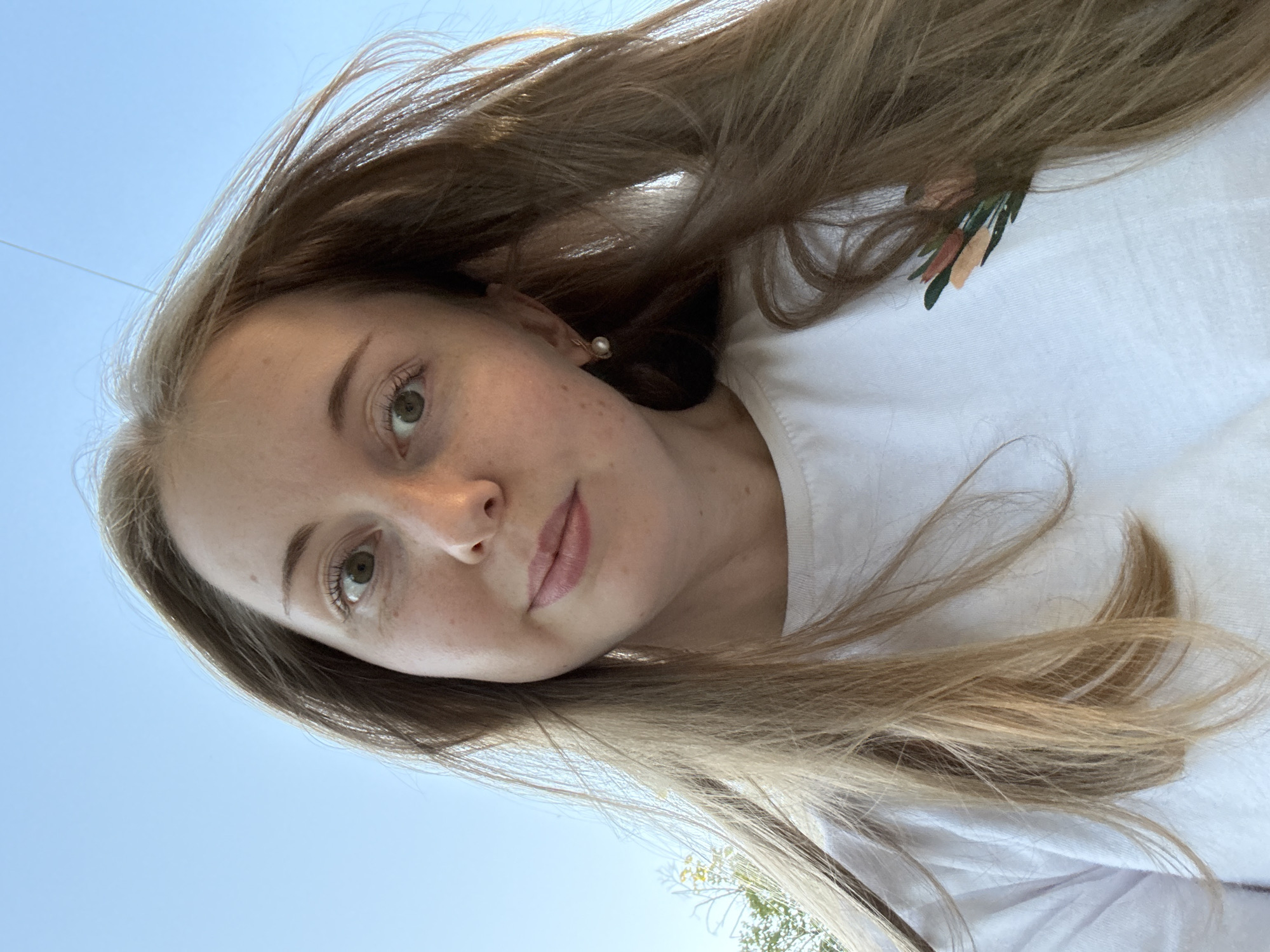 A young woman with long blonde hair is looking at the camera in a close-up, selfie-style shot. She is wearing a white t-shirt with a small floral design on the left side of her chest. The background is a clear blue sky. She has green eyes and is wearing a pearl earring on her left ear. The lighting suggests it is daytime, possibly late afternoon due to the warm tones. The environment appears to be outdoors, with a hint of greenery visible on the far left edge of the image. The focus is on her face and upper body.