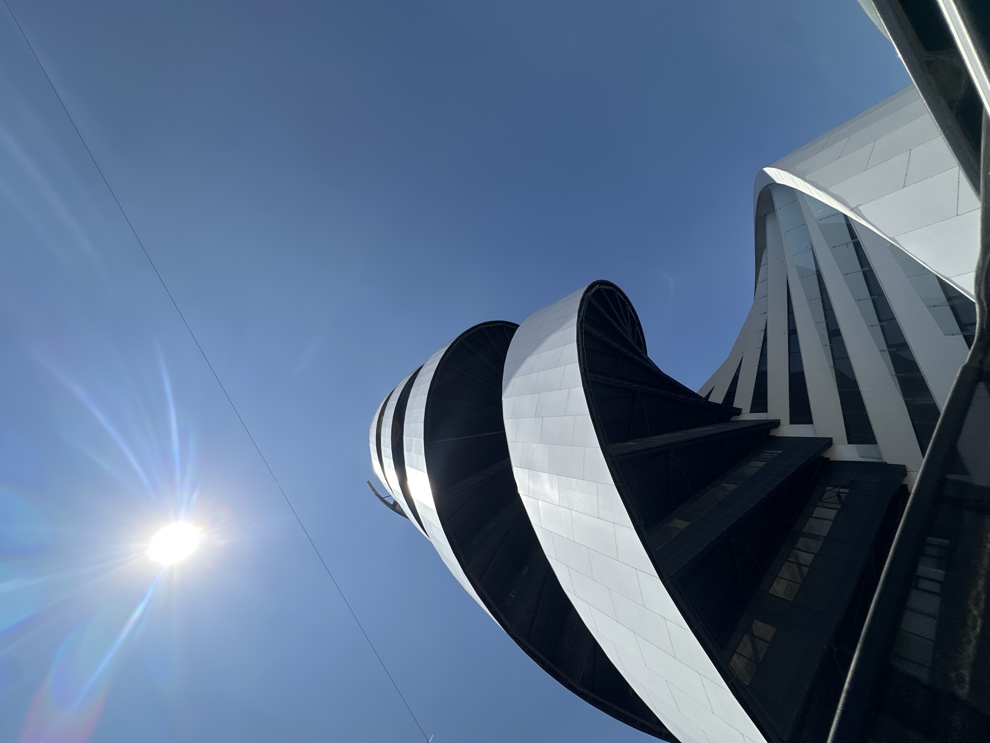 The image displays a modern, spiraling architectural structure in San Bernardino Tlaxcalancingo, Mexico, viewed from a low angle against a clear, bright blue sky. The building exhibits a helical design, characterized by continuous bands of white paneling that curve and ascend, alternating with darker, recessed levels that feature horizontal window arrays. The sun is prominently visible in the upper left quadrant, emitting intense light and distinct lens flares exhibiting spectral colors. A single, dark, thin cable spans horizontally across the upper portion of the sky, below the sun. A dark, out-of-focus barrier or railing is present in the immediate lower foreground. No individuals or overt activities are discernible within the frame.