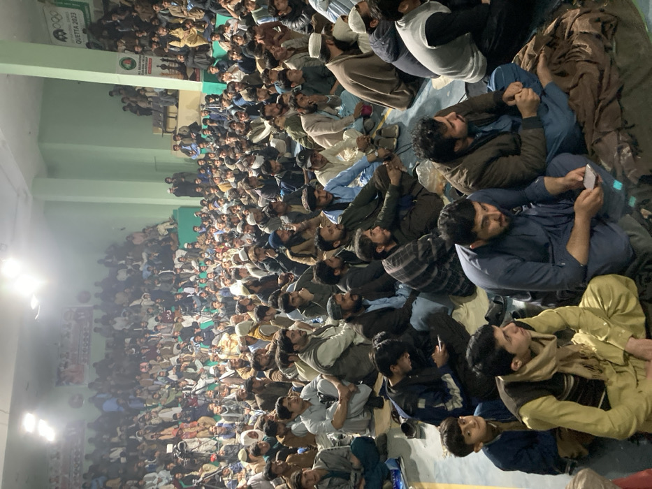 An indoor, wide-angle shot captures a large gathering of predominantly male individuals in Quetta City Tehsil, Pakistan, within a spacious hall. The attendees, numbering in the hundreds and ranging from children to adults, are seated closely together on the floor and on elevated bleachers/platforms. Most are dressed in traditional South Asian attire, including shalwar kameez and waistcoats, with some wearing skullcaps or other head coverings. The room features light green walls and is illuminated by bright overhead lights.
In the background, two banners are visible on the far wall: one displaying "QUETTA 2023" alongside a logo resembling the Olympic rings, and another partially obscured banner that reads "BALOCHISTAN CHAMPIONSHIP." Several individuals are seen operating professional camera equipment mounted on tripods among the crowd on the left side of the frame, indicating media coverage.
The crowd generally faces forward, suggesting an event or presentation. Individuals in the foreground are seated on what appear to be blankets or mats. Some are observed looking towards the camera, while others maintain their focus forward. At least two individuals in the foreground are holding mobile phones, one actively engaged with the device. No direct interactions between individuals are prominently displayed beyond shared proximity. The overall environment suggests a public assembly or community event.