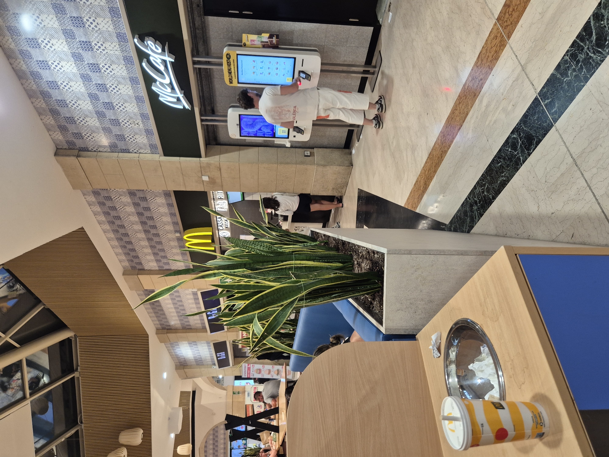 The image depicts the interior of a McDonald's restaurant with a McCafe section in Taranto, Italy. In the foreground, a wooden counter displays a disposable cup with a straw and a stainless steel bin filled with used paper napkins. In the midground, a large planter box contains several snake plants. To the right, two self-service kiosks are visible, with one customer interacting with the touchscreen. In the background, other patrons are seated at tables, and a McDonald's logo is prominently displayed above the McCafe counter. The restaurant features a checkered tile pattern on the walls and a ceiling with wooden slats and a skylight. The floor is a polished marble with inlaid bands of different colors.