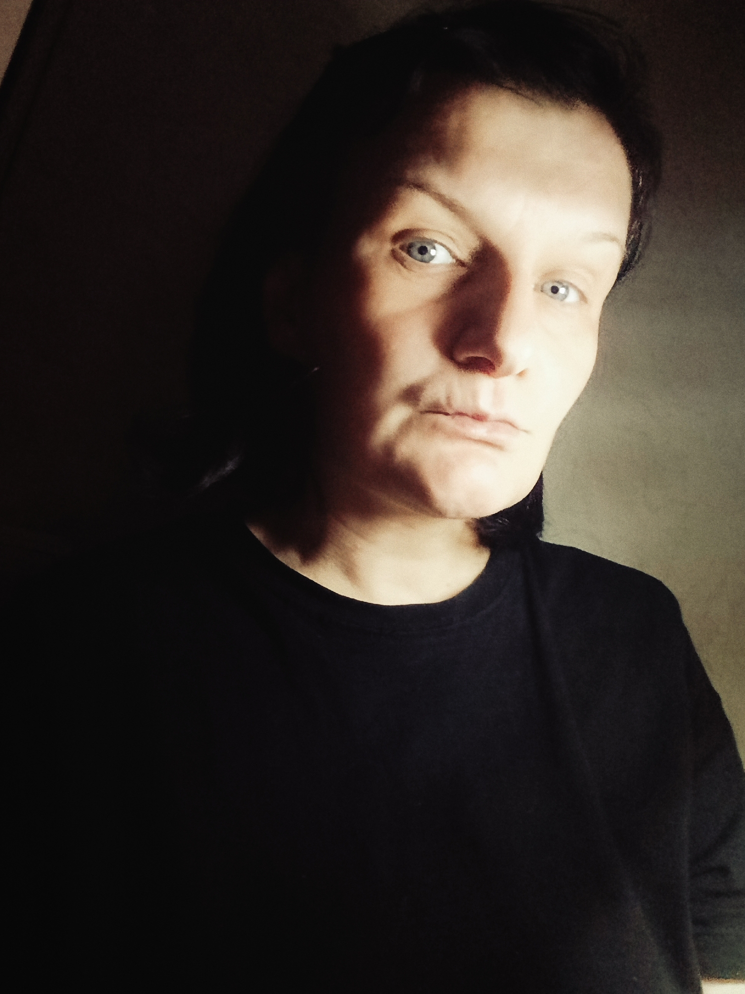 A headshot-style portrait features an adult person, likely female, looking directly into the camera. The individual has light skin, prominent blue eyes, and dark hair that appears to be short or pulled back, framing their face. They are wearing a plain, dark-colored, crew-neck t-shirt.
The lighting is dramatic and high-contrast, illuminating the right side of the face and upper chest while the left side is cast in deep shadow, creating a chiaroscuro effect. The background is simple and indistinct, appearing dark on the left and a lighter, muted tone on the right, suggesting an indoor setting. The person holds a neutral to slightly serious expression, with their lips gently closed. No specific activity is depicted beyond the act of posing for the photograph. There is no visible text in the media file. The image is associated with the location Фоки, Russia.