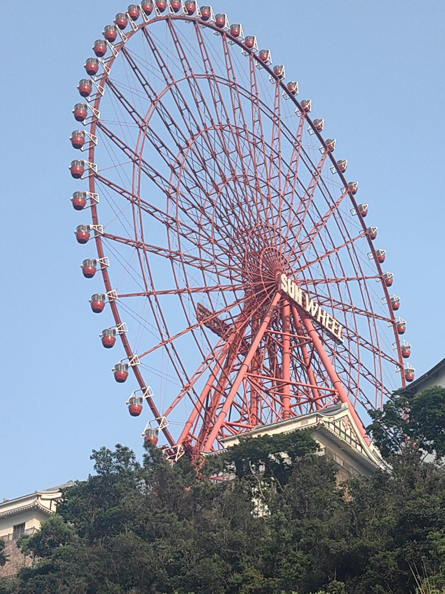 This vertical shot captures a large, vibrant red Ferris wheel set against a clear, light blue sky. The Ferris wheel dominates the frame, showcasing its intricate lattice work and numerous red passenger cabins distributed along its circumference. In prominent white capital letters, the words "SUN WHEEL" are visible on the central hub of the structure.
Below the Ferris wheel, a dense canopy of green trees and foliage partially obscures a building. This building exhibits distinct traditional Asian architectural features, including an ornate, multi-tiered curved roof with decorative gables.
The scene appears to be an amusement or tourist area during the daytime, under bright and clear weather conditions. No people or specific activities are discernable, but the presence of the Ferris wheel suggests a leisurely and entertaining environment. Based on the provided context, the location is Hong Gai Ward, Vietnam.