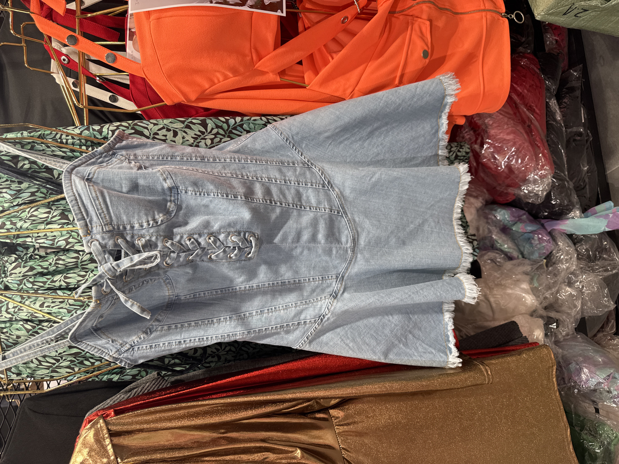 This is a close-up, eye-level, interior shot of a retail clothing rack in Gungoren, Turkey. The rack displays several garments, including a light blue denim corset dress with lace-up detailing and a frayed hem, a metallic gold long-sleeved garment on the left, a green patterned dress in the background, and a neon orange jacket or top on the right. Beneath the hanging garments, several items are visible in plastic packaging, appearing to be folded clothing. The scene suggests a commercial environment, likely a clothing store or market stall. No people are visible in the frame.
