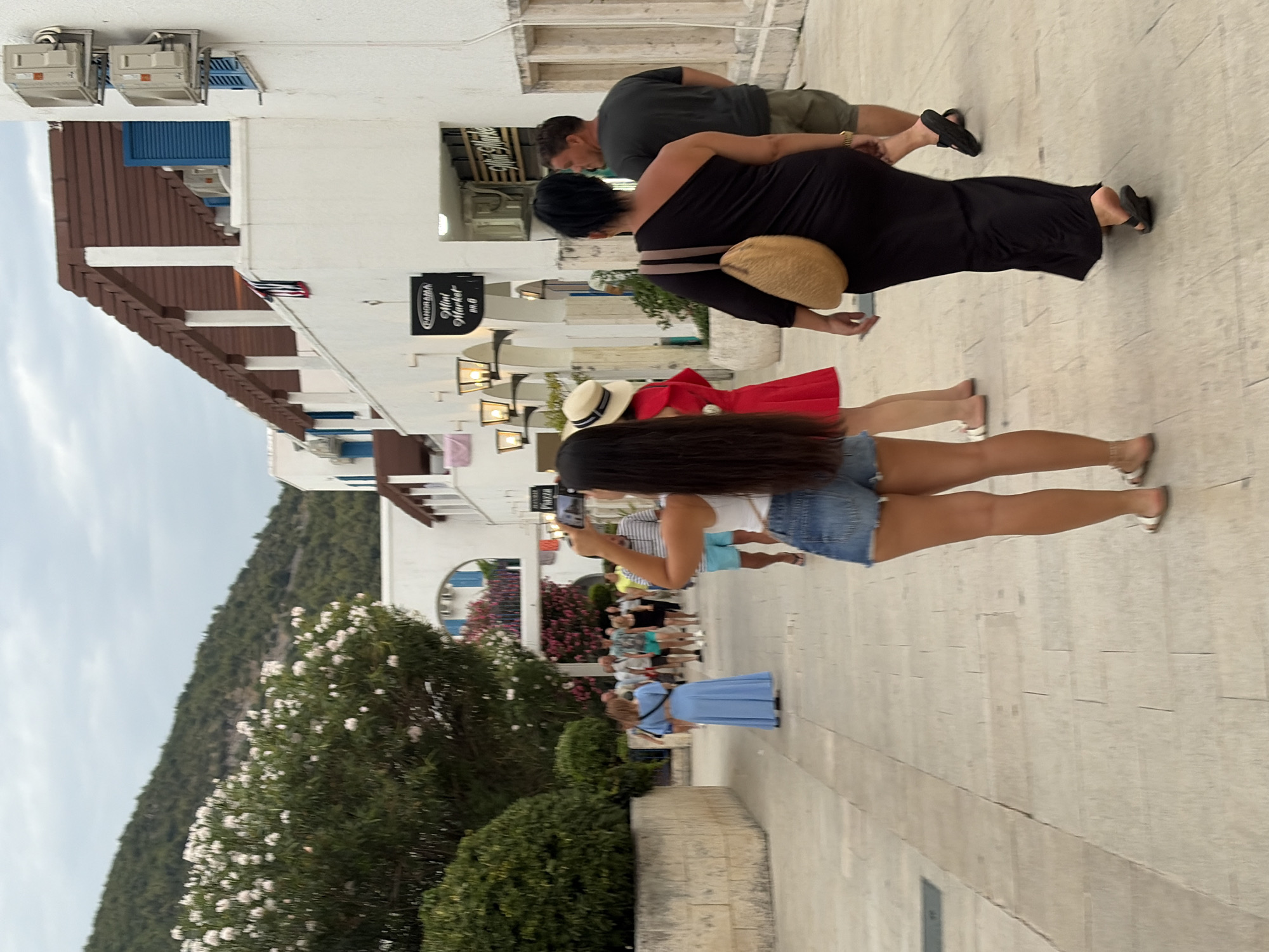 The image depicts a street scene in Budva, Montenegro, with several individuals walking along a paved pathway lined with white buildings. In the foreground, a woman with long dark hair is taking a photo with her phone. She is wearing denim shorts and a white top. To her left, a woman in a long blue dress walks away from the camera. Further down the path, a group of people are walking in the same direction. One person is wearing a red dress and a straw hat. On the right side of the image, a woman in a black one-shoulder maxi dress with a straw bag slung over her shoulder walks away from the camera. Beside her, a man in a grey t-shirt and shorts is walking. The buildings have a Mediterranean architectural style, with white facades, wooden pergolas, and blue shutters. Lush green trees and flowering bushes are visible on the left side of the image, and a hillside covered in vegetation forms the background. The sky is overcast. Signs for "Panorama Mini Market" are visible on one of the buildings.