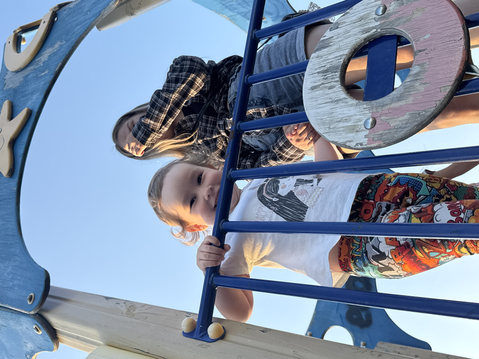 A low-angle shot captures a young child and an adult on a playground structure. The child, wearing a white t-shirt with a graphic design and comic-style shorts, smiles towards the camera while holding onto the blue metal bars of the play structure. Behind and above the child, an adult in a black and white checkered shirt and grey shorts is visible. A round, worn wooden component with visible screw heads is near the adult. The playground appears to be painted in blue and grey colors. The background is a clear, bright sky.
I can not determine the location in the city of Фоки, Russia from the image.