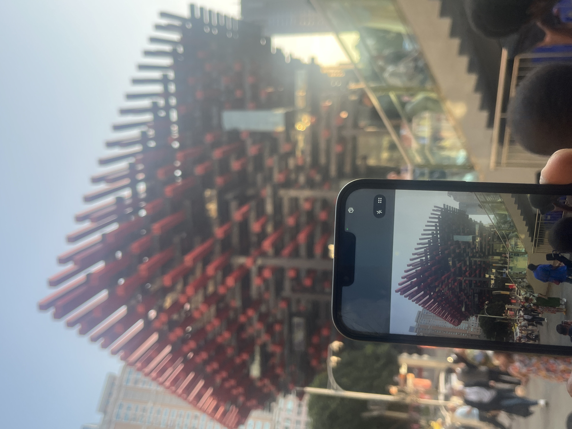 A slightly blurred, medium-angle outdoor daytime shot shows a person holding a smartphone to take a picture of the "Liziba Station Pier" in Yuzhong District, China. The station, adorned with a chaotic but organized mass of red planks, occupies much of the left side of the image, towering above the surroundings.
The smartphone in the foreground captures a similar view, but from a lower angle, showing people walking below the elevated train station. The surrounding environment appears urban, with buildings, trees, and streetlights visible. People are also walking behind the person taking the photograph. The sky is a pale blue, slightly obscured by the sun’s glare at the top of the photograph.