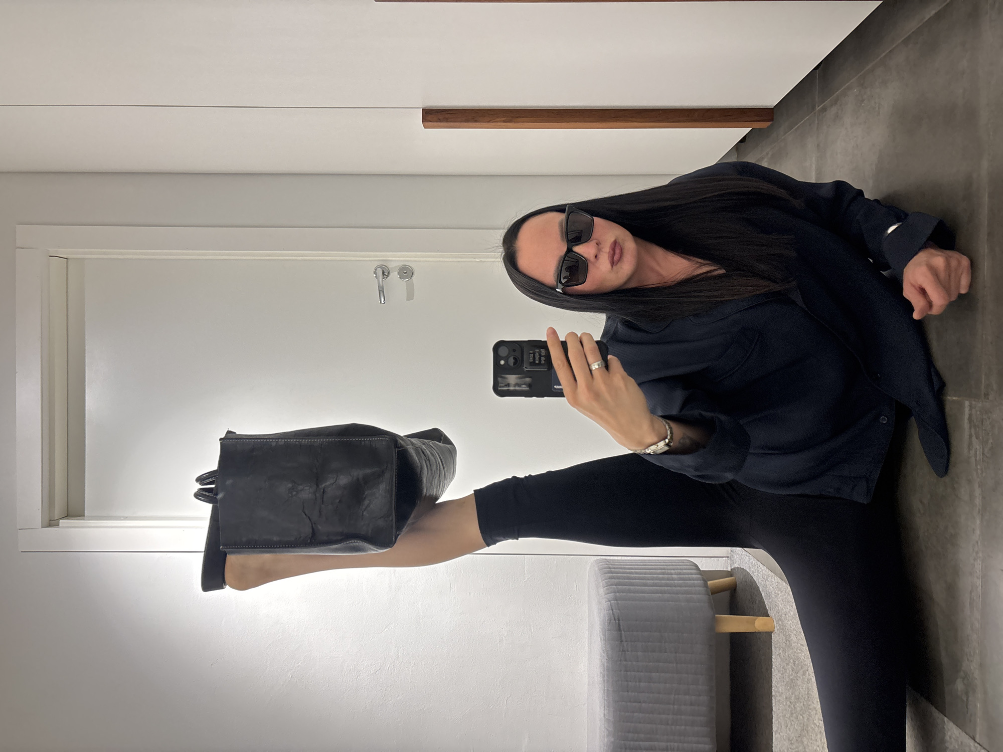 A woman with long dark hair is posing for a selfie in what appears to be a modern interior, possibly a closet or dressing room. She is wearing sunglasses, a dark button-down shirt, and black leggings. She has one leg extended upwards, holding a black handbag by the ankle. She is holding a black smartphone in her right hand, taking the photo. Behind her, there is a white door with a silver handle and a gray ottoman with wooden legs. The flooring is gray, and there is a white cabinet with a wooden accent in the background. The image does not contain enough information to determine if it was taken in Chaikovsky, Russia.