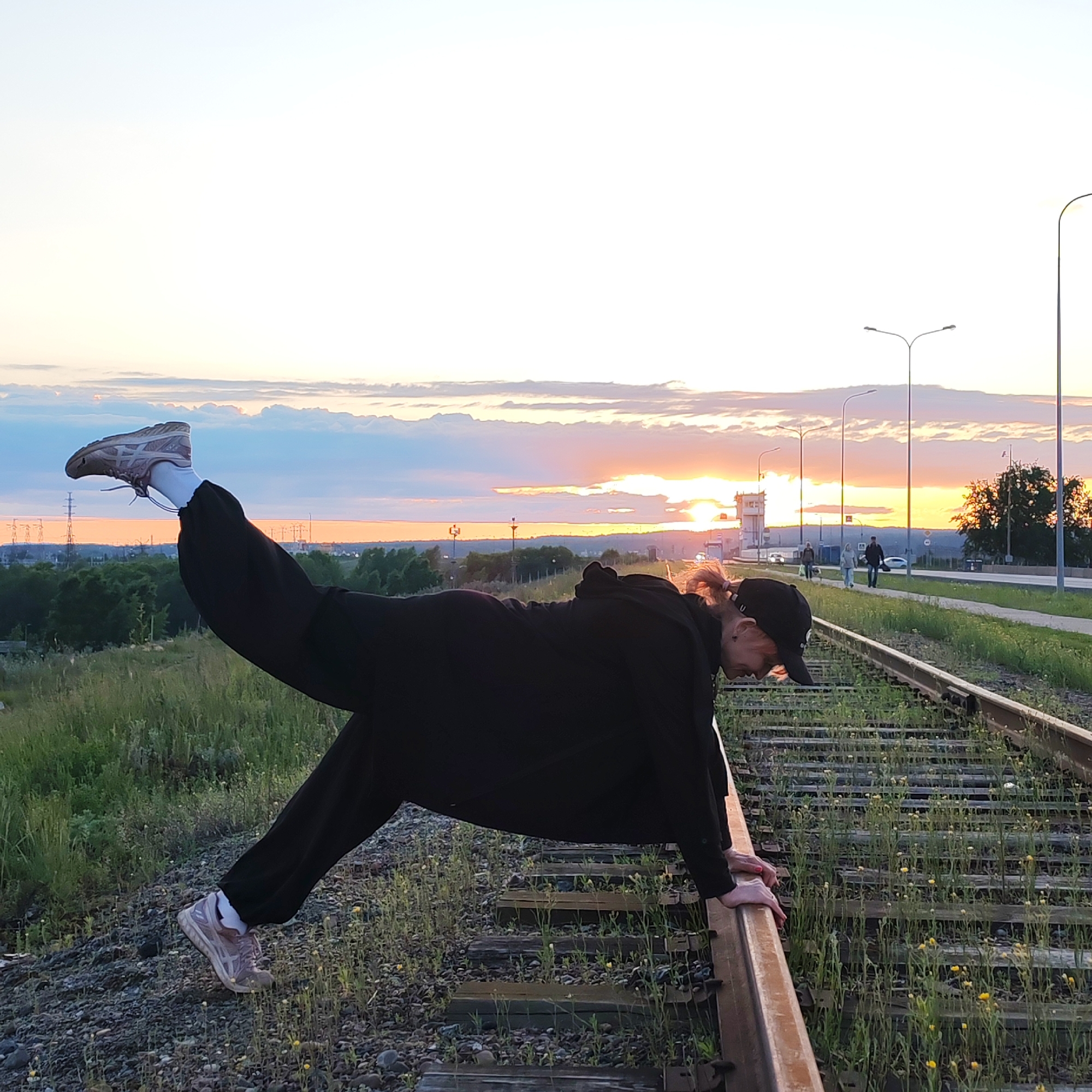 The image depicts a person performing an athletic or artistic pose on overgrown railway tracks during sunset in Chaikovsky, Russia. The subject, a young individual, is dressed in a black tracksuit, a black baseball cap, and light-colored sneakers with white socks. They are captured mid-pose, holding onto one of the rusted railway tracks with both hands, their body extended horizontally, and their left leg raised high in the air, creating a striking silhouette against the sky. Red nail polish is visible on the person's hands.
The railway tracks are partially covered with green grass and small yellow flowers, indicating they may be disused or infrequently used. An grassy embankment slopes upwards to the left. In the background, a vivid sunset bathes the sky in warm oranges, pinks, and yellows, fading into blue and white clouds above. The sun is visible on the horizon, casting a soft, golden light across the scene.
Parallel to the tracks, a road is visible, lined with modern streetlights. Distant figures can be seen walking along the road, and several vehicles are present. Further in the background, a mix of trees, power lines, and a distinctive white building (possibly a control tower) against the horizon suggests a semi-urban or industrial landscape. The time of day is clearly evening, characterized by the dramatic sunset. No text is visible in the image.