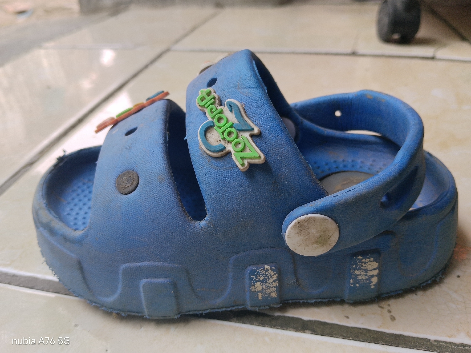 A blue children's clog is photographed from a side profile, resting on a tiled floor. The clog features a strap across the top and a decorative charm with green and blue lettering. The shoe appears worn and dusty, with scuff marks visible on its surface. The background is blurred but shows additional tiles and a dark, out-of-focus object. The image is a close-up of the footwear, with a watermark indicating "nubia A76 5G" in the bottom left corner. This photograph was taken in Binuang, Indonesia.