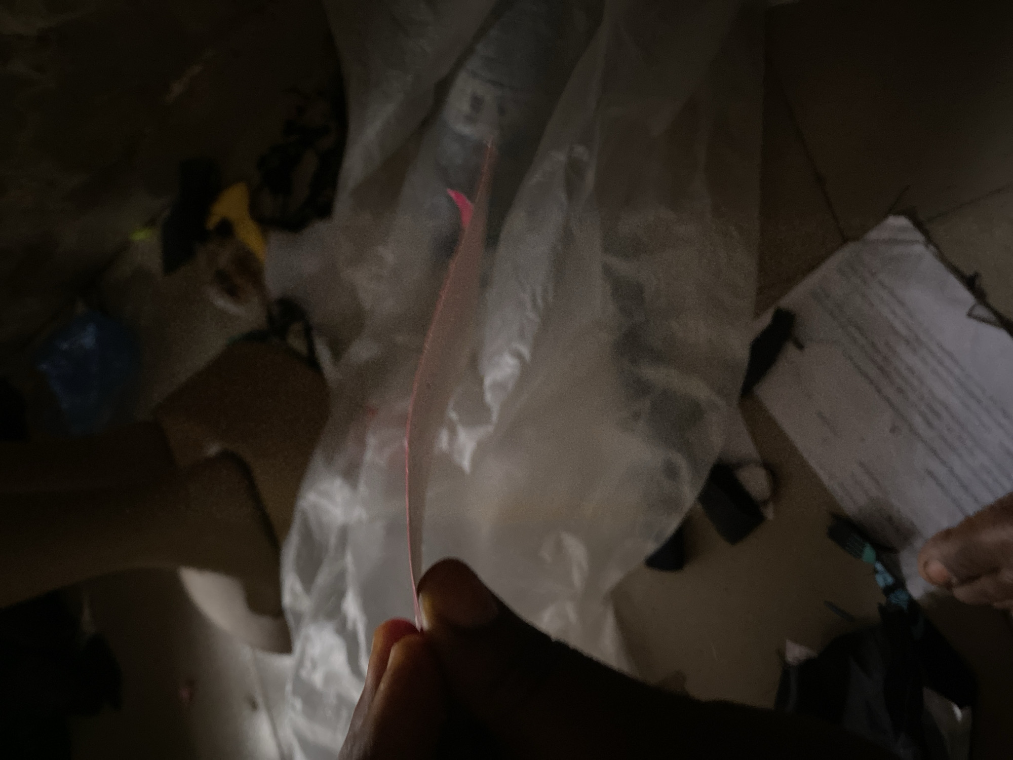 The image depicts a close-up view of a hand holding a pink, thin object, possibly a piece of paper or plastic, near a large, translucent plastic bag. The background is cluttered with various items, including what appears to be a mannequin leg, fabric scraps, and other unidentifiable objects, all in a dimly lit environment. The scene suggests a disorganized, indoor space, potentially a storage area or a workshop. The location of Jauro Yinu, Nigeria, is not visually evident in the image itself; it is contextual information provided separately.