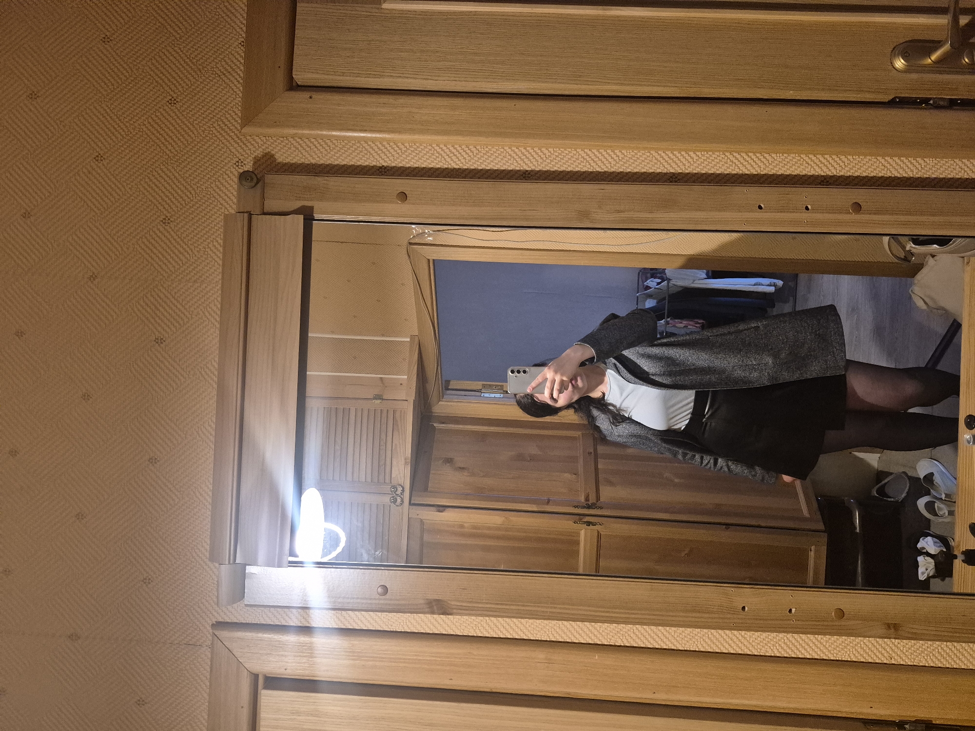 The image shows a full-length mirror reflecting a person standing in what appears to be a bedroom. The person is holding a smartphone and taking a selfie. They are wearing a grey blazer over a light blue top and a black skirt, with black tights. In the background of the reflection, there are wooden wardrobes and cabinets. To the right of the reflection, shoes and other items are visible on the floor, suggesting a dressing area. The room has patterned wallpaper on the walls. The lighting suggests it might be evening or indoors with artificial light, as there is a bright light source on the left side of the mirror. The overall atmosphere is casual and personal, capturing a moment of self-reflection.