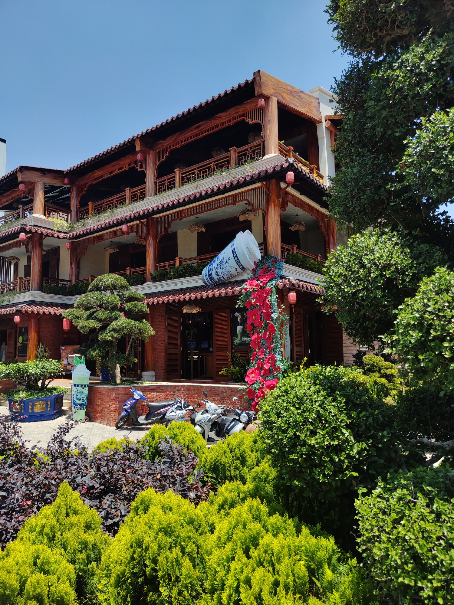 The image shows a three-story building with traditional Vietnamese architectural features located in Cam Ly - Đà Lạt, Vietnam. The building is constructed primarily of wood, with red tiled roofs, and decorative lanterns. A large, sculpted replica of a drink container is attached to the building’s upper level, appearing to “pour” a cascade of red flowers down the facade. In front of the building, there are three parked motorbikes, and the foreground is filled with various shrubs and plants. The sky is clear blue.