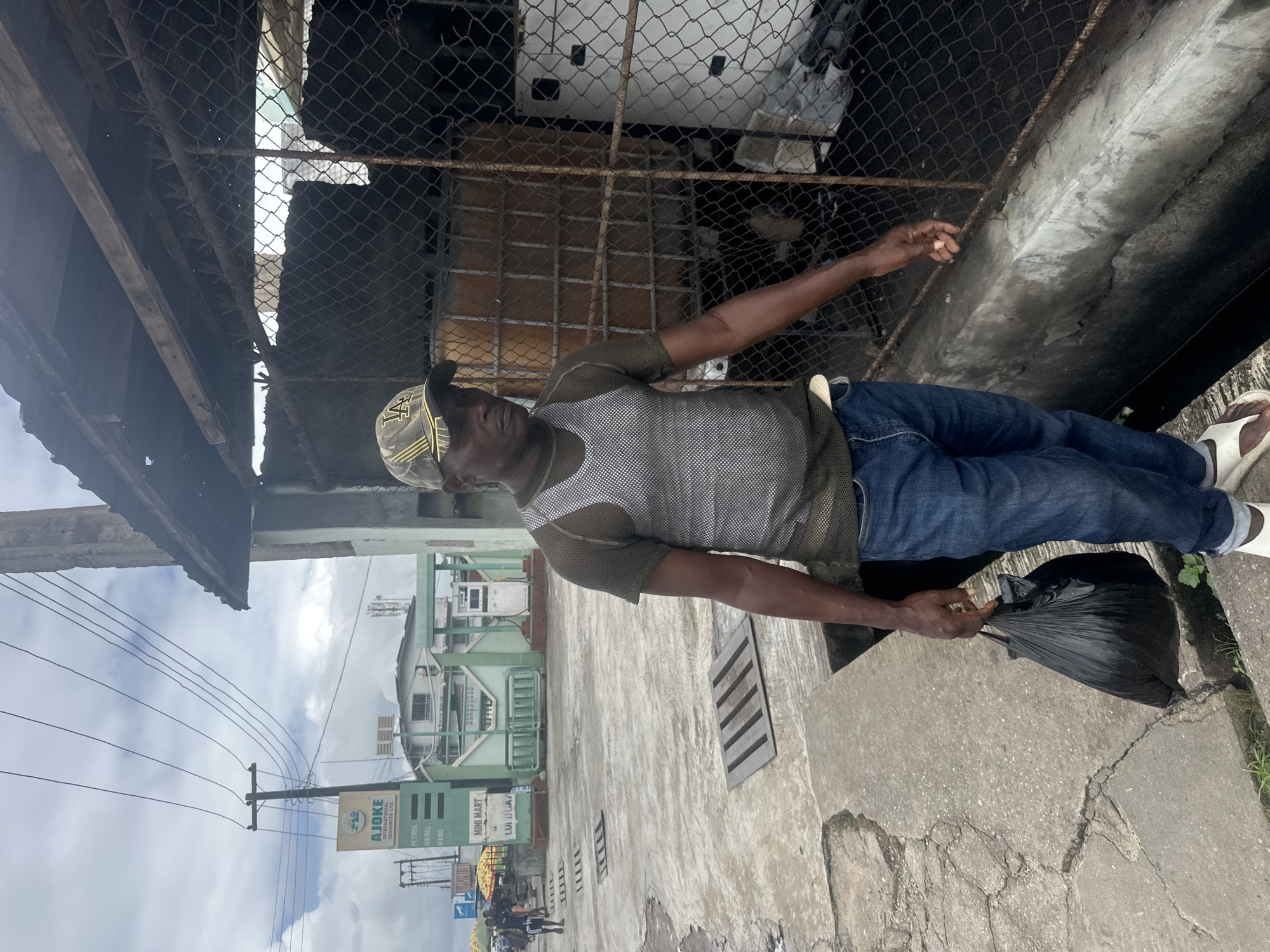 A Black man wearing a camouflage cap, a grey sleeveless shirt over a dark t-shirt, and blue jeans walks along a concrete pathway holding a black plastic bag. He is in an outdoor urban environment with a cloudy sky. In the background, a green and white building with "AJOKE" signage and fuel pumps is visible, suggesting a gas station or convenience store. A wire fence encloses an area with large containers and a white appliance. The pathway is cracked and has metal grates. The man's expression is neutral as he looks forward. The overall scene depicts a street in Badagry, Nigeria, likely near a commercial establishment.