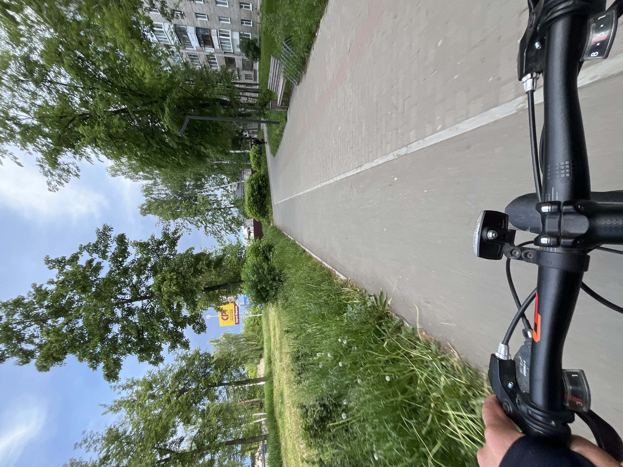 A low-angle shot taken from the perspective of someone riding a bicycle on a paved path. The bike's handlebars and front light are visible in the foreground. The path is bordered by grass and trees on one side and a building on the other. A sign is visible among the trees, bearing the logo "CP" and the text "Россия". The location is in the city of Chaikovsky, Russia. The sky is partly cloudy.