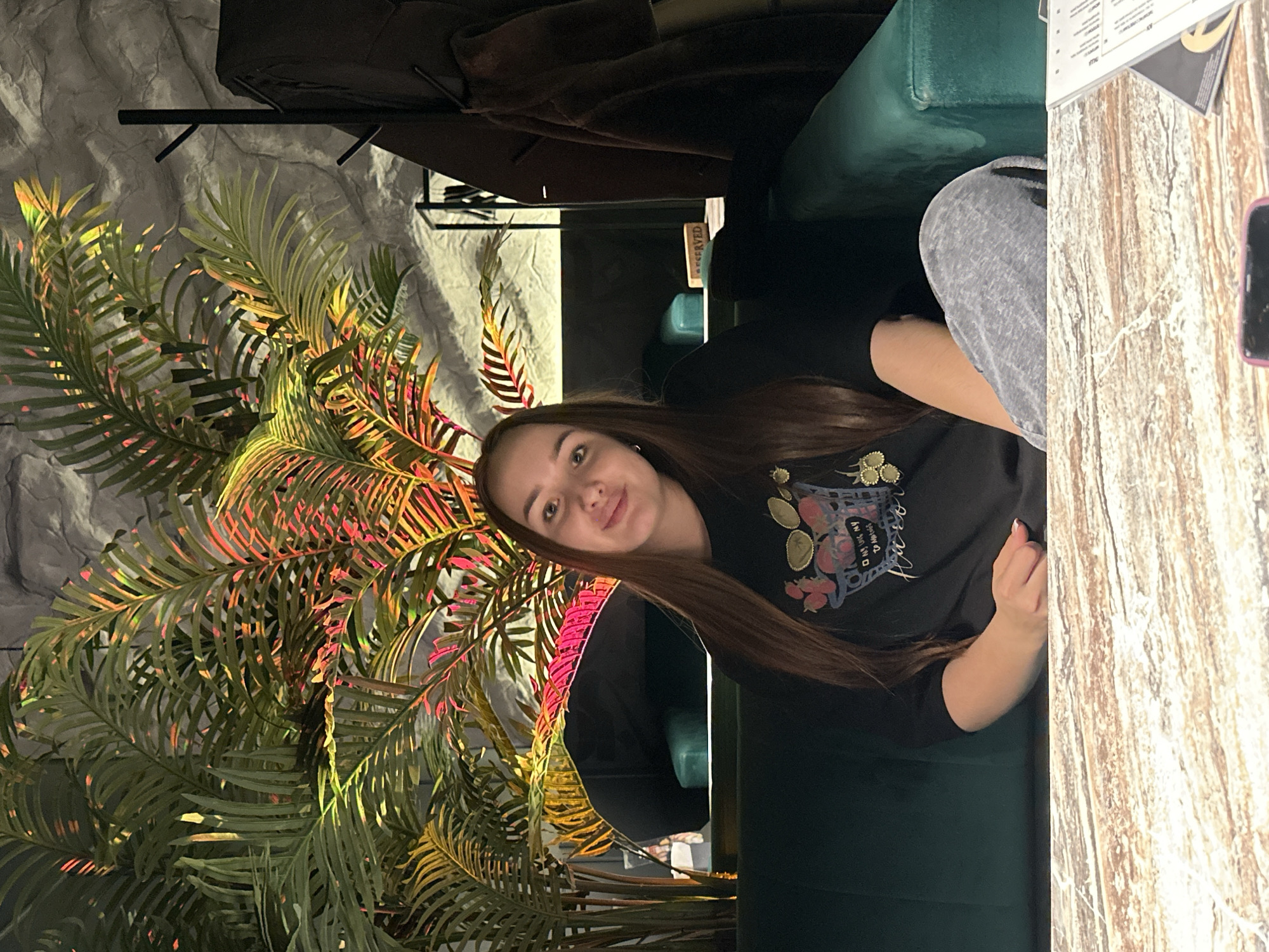 A young woman with long brown hair is seated at a table in what appears to be a restaurant or cafe, likely located in Chaikovsky, Russia. She is wearing a black long-sleeved shirt with a graphic design. The table has a faux wood finish, and there is a menu and a mobile phone on it. The woman is smiling and appears to be posing for the camera. Behind her, there's a green velvet booth or chair. The background includes a wall with a textured, rock-like appearance, as well as a large artificial palm tree illuminated with colored lights, giving the scene a tropical or exotic ambiance.