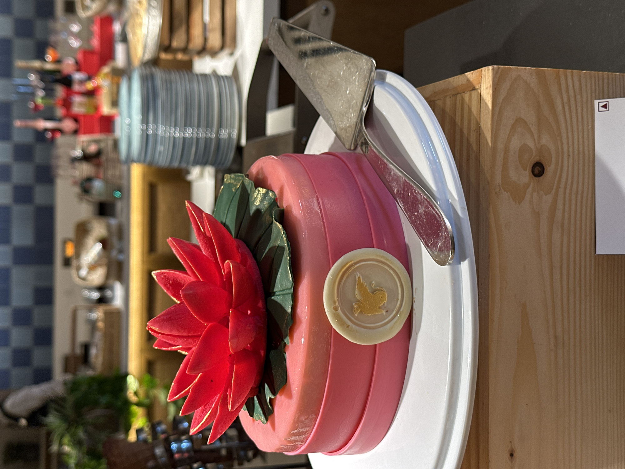 A bright pink cake with a red lotus flower decoration sits on a white plate, which is placed on a wooden surface. A silver cake server rests on the plate, partially in front of the cake. The cake has a gold emblem with a bird on its side. In the background, there are stacked blue plates and a blurry scene of a buffet. The image appears to be taken indoors, possibly in a restaurant or hotel in Vagator, India.