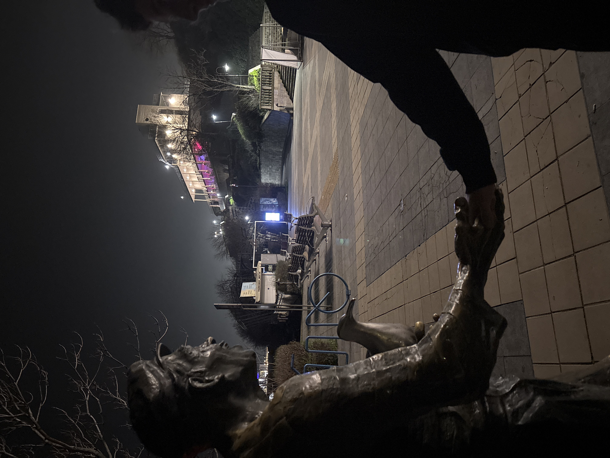 At night in Burgas, Bulgaria, a bronze sculpture of a reclining figure is situated on a brick-paved promenade. The sculpture's hand is being grasped by a person wearing a black garment. In the background, there's a multi-story building with illumination and architectural details, possibly a hotel or civic structure. Bare branches of trees are visible against the night sky. Streetlights are present, casting light onto the scene. There are benches and bicycle racks in the middle distance.
