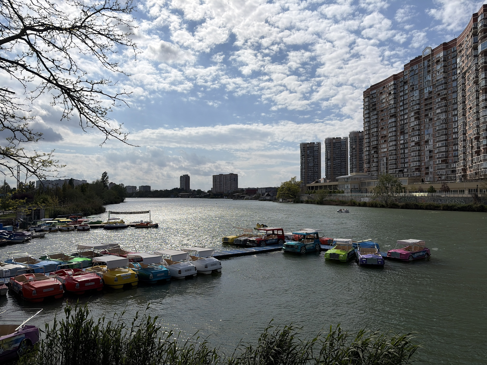 The image captures a waterfront scene in Krasnodar, Russia, featuring a body of water with several brightly colored pedal boats docked and floating. The boats are designed to resemble vintage cars and are grouped along docks and in the water. In the background, there are clusters of tall apartment buildings and other urban structures, indicating a city environment. The sky is partially cloudy with sunlight reflecting on the water's surface. Some trees are visible along the shore. The vegetation in the foreground consists of tall grasses and shrubs.