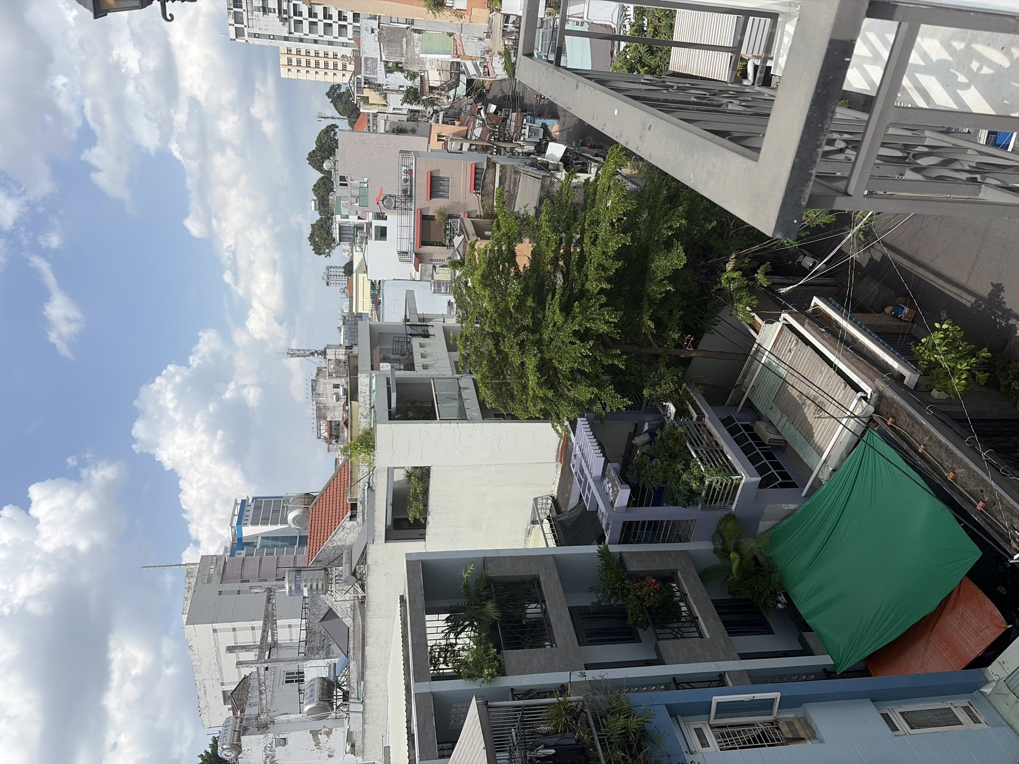 This high-angle, vertical shot captures a dense urban residential landscape in Thủ Đức, Vietnam, under a bright, partly cloudy sky during the daytime. The scene is observed from an elevated vantage point, likely a balcony or rooftop, indicated by a dark metal railing in the foreground.
The area is characterized by tightly packed, multi-story buildings of various designs and ages, common in Vietnamese cities. Many structures feature balconies adorned with plants, exterior air conditioning units, and rooftop water tanks. A narrow street or alley runs through the middle ground, lined by buildings and crisscrossed by overhead power lines. A large, prominent green tree stands in the mid-ground, providing a burst of natural color amidst the concrete, and other smaller trees and greenery are scattered throughout the scene.
Below the vantage point, a large green tarp or awning covers an area, with a smaller reddish-brown tarp visible nearby. No people are visible, and no specific activities are taking place, suggesting a quiet moment in the urban environment. The distant background reveals taller, more modern high-rise buildings under a clear blue sky, contrasting with the older, lower-rise residential structures. The overall impression is one of a bustling, lived-in urban area with a blend of architecture and green spaces. No discernible text is visible on any signs or buildings.