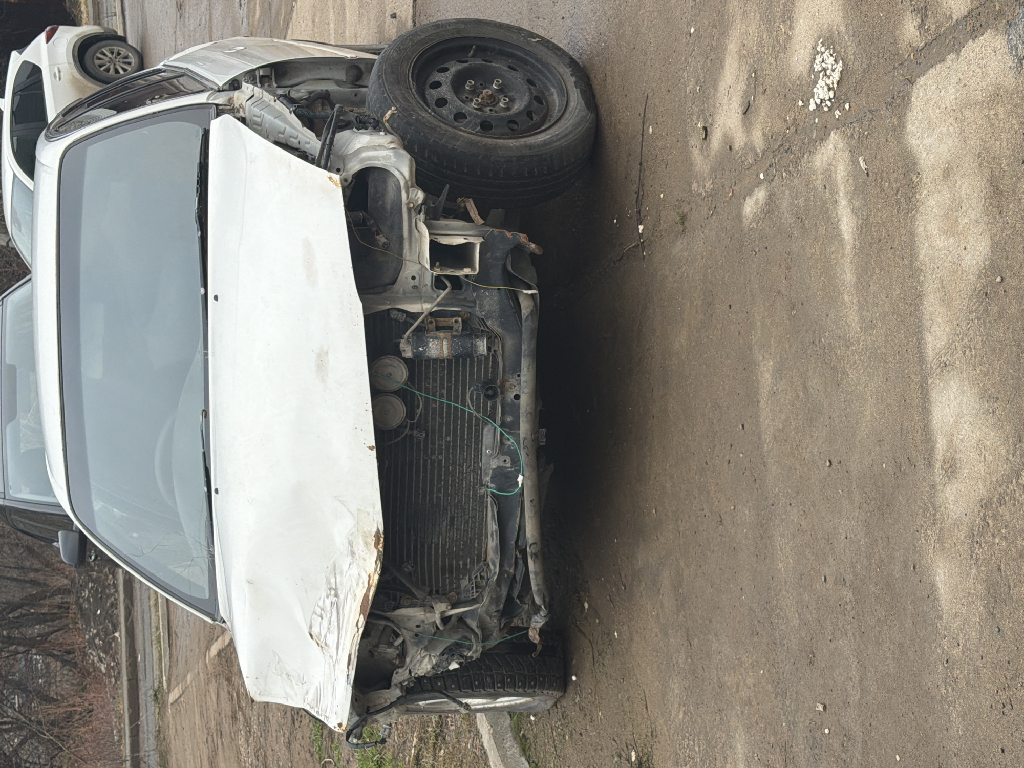 The image shows a severely damaged white car on a paved surface, likely a road or parking lot. The car's front end is heavily crumpled, with the hood significantly deformed. A tire is detached and resting on the front portion of the vehicle. The radiator is exposed and damaged. The environment appears to be an outdoor urban setting. The location is Chaikovsky, Russia. Other vehicles can be seen parked nearby.