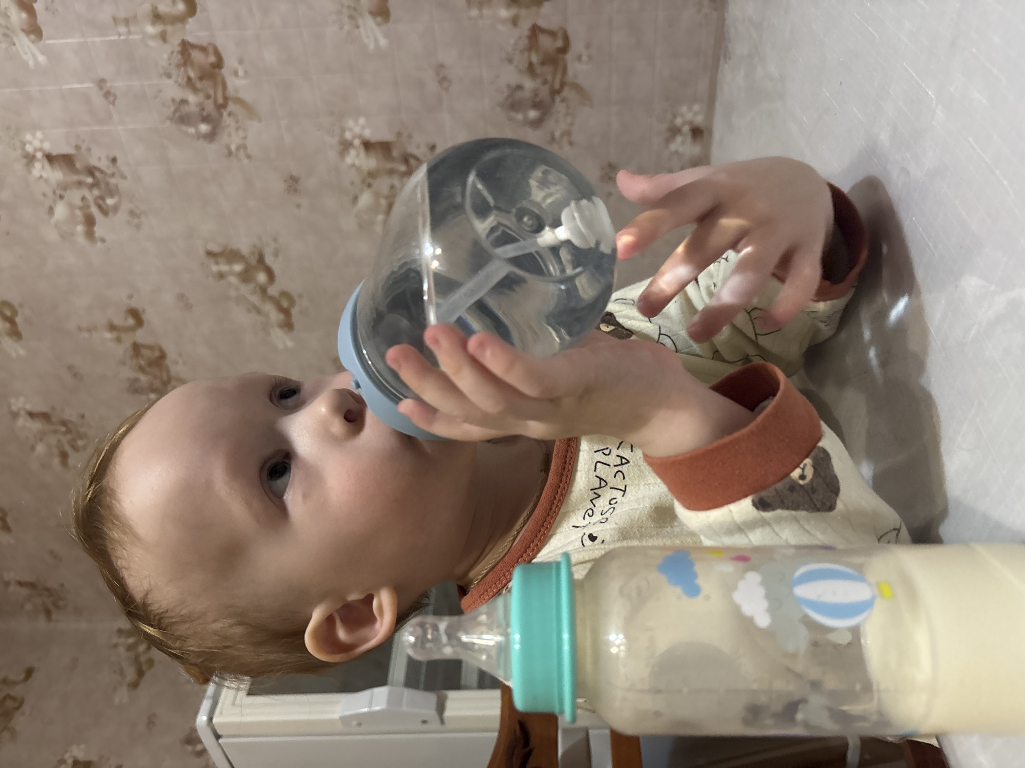 The image displays a young child, likely a baby or toddler, lying horizontally in a brightly lit indoor setting. The child has fair skin, light blonde or light brown hair, and blue eyes. They are focused on drinking from a clear plastic bottle with a light blue cap and a straw-like mechanism inside, held with both hands. The bottle appears to contain clear liquid or is partially empty.
The child is wearing a long-sleeved onesie or top. The main fabric is a light cream or beige color, adorned with small, dark, possibly cartoonish patterns. The cuffs at the wrists and the trim around the collar are a contrasting rust or orange-brown color. On the left shoulder/chest area of the clothing, the words "CACTUS PLANET :)" are visible, written vertically. A small illustration of a brown bear with a white muzzle is also present on one of the arm cuffs.
To the right side of the frame, near the child's lower body, another baby bottle is visible. This bottle is filled with a milky white liquid, likely formula or milk. It has a teal or turquoise cap and nipple, and is decorated with light blue and white clouds, and a blue and white striped hot air balloon.
The background consists of two distinct areas. Behind the child's head and upper body is a wall covered with patterned wallpaper. The wallpaper features light brown or beige floral and abstract designs on a slightly lighter beige background. To the right of the child and extending towards the bottom of the frame is a plain, light-colored surface, possibly a wall or a piece of furniture, which contrasts with the patterned wallpaper. A small section of a white and brown object, possibly furniture, is also visible in the lower left corner of the image. The overall lighting suggests an interior, possibly during the daytime. The child's expression is neutral and concentrated on drinking.