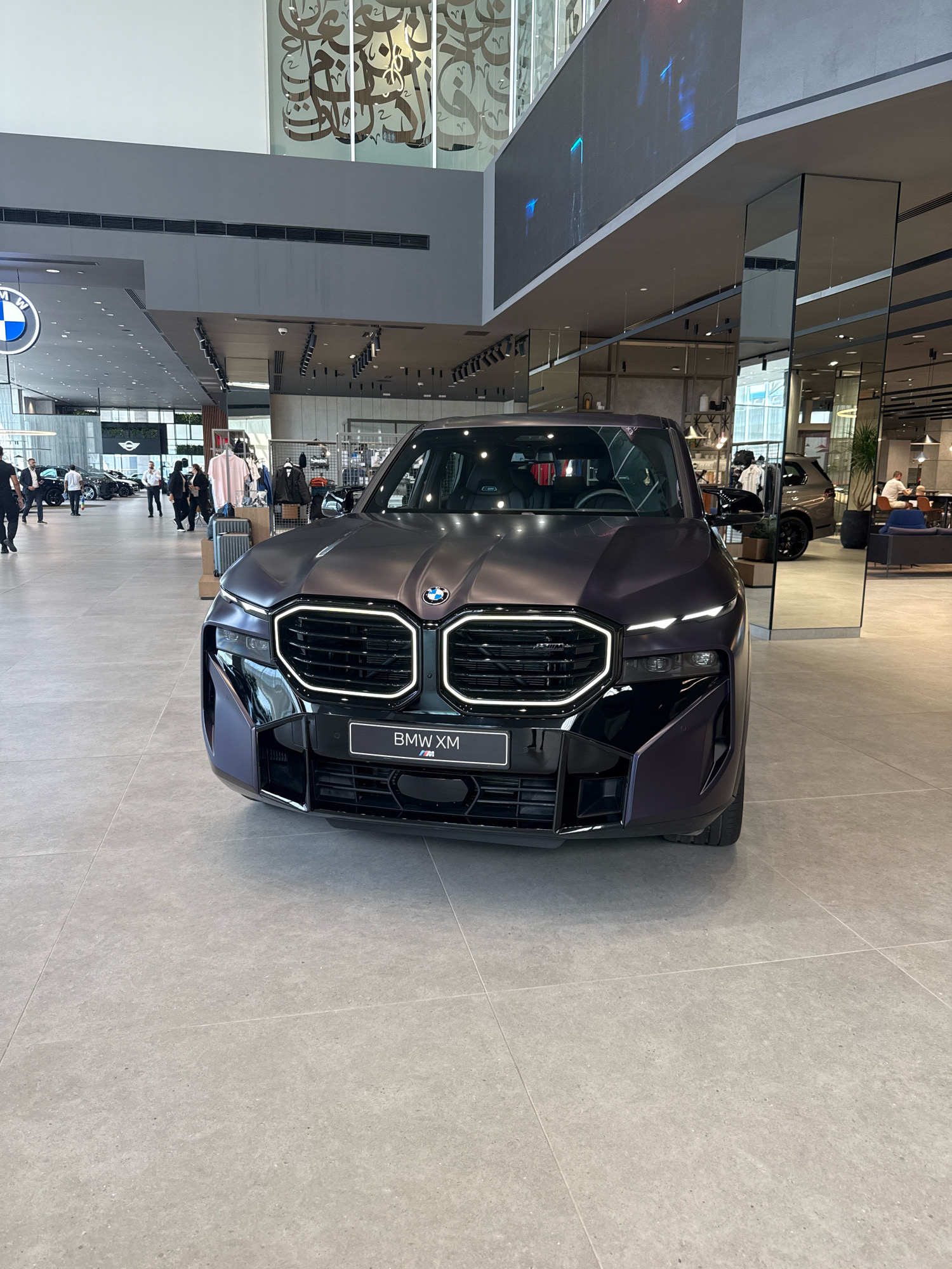 The image shows the front of a dark purple BMW XM parked indoors, likely in a car dealership. The car is the central focus, with its distinctive grille and headlights illuminated. Behind the car, the environment appears to be a modern showroom with polished tile flooring, large windows, and overhead lighting. In the background, there are other cars on display, people walking around, and merchandise racks with clothing. Reflections in large mirrored panels suggest a spacious and upscale setting. On the wall above, there is a large piece of artwork with Arabic calligraphy. The overall impression is a contemporary commercial space designed to showcase luxury vehicles. The location is identified as Dubai, United Arab Emirates.