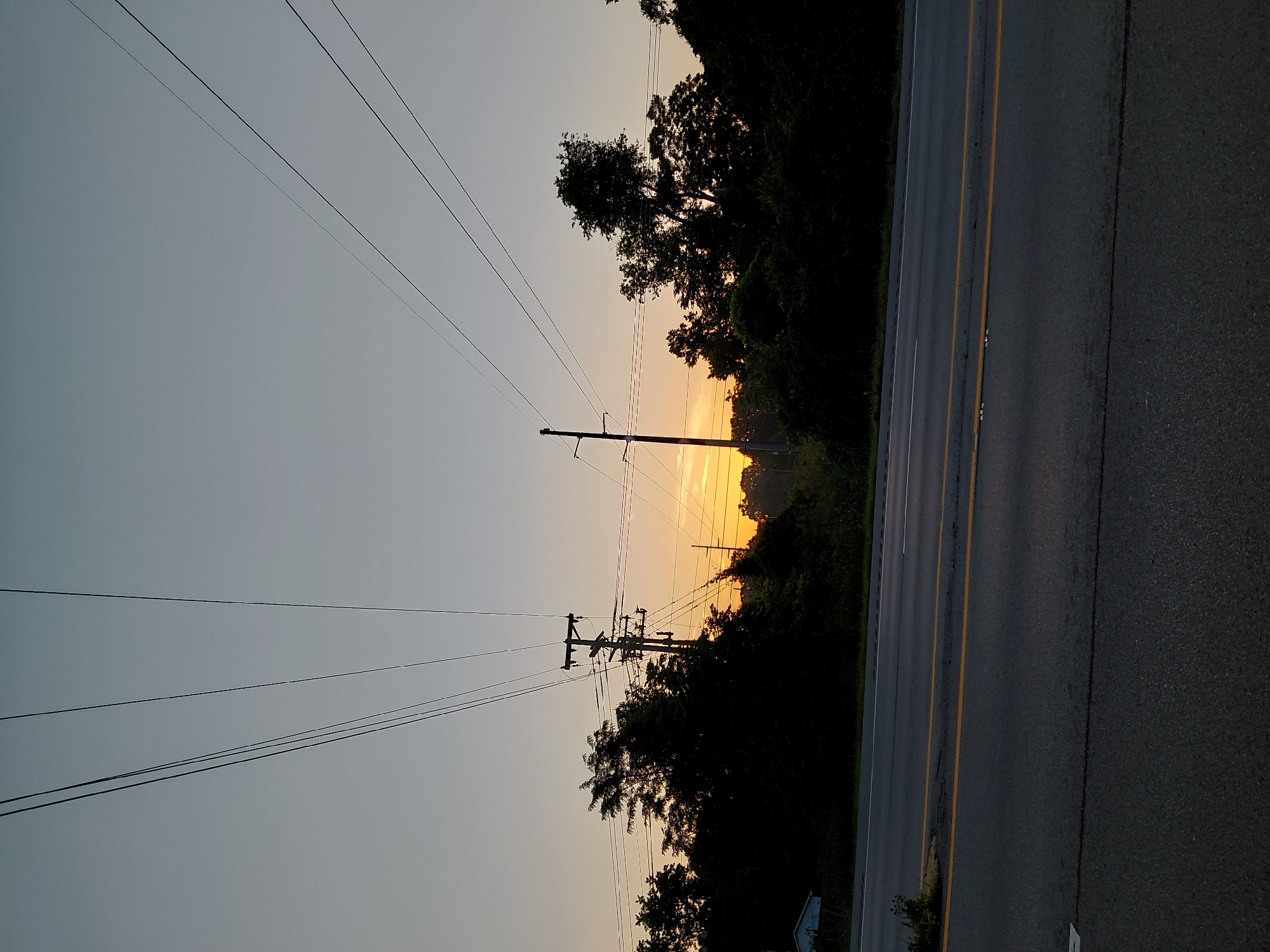 The image captures a view of a road and power lines against a dusky sky, likely at sunrise or sunset. The sky is a pale gradient of grey and orange, with the sun's glow visible near the horizon, partially obscured by silhouetted trees and utility poles. Multiple power lines stretch across the frame, originating from two prominent utility poles. One pole is situated on the right side of the image, with the other slightly to the left, closer to the center. Dense, dark foliage and trees form a thick line along the horizon, obscuring the ground behind them. In the far left background, a small, light-colored building, possibly a shed or small house, is partially visible through the trees. The foreground shows an asphalt road with yellow and white lane markings. The road appears to be a multi-lane highway. The scene is devoid of people or any discernible activity. The overall impression is a quiet, somewhat desolate roadside landscape during twilight. The location is not specified in the image itself and therefore cannot be identified as Frankfort, United States of America.