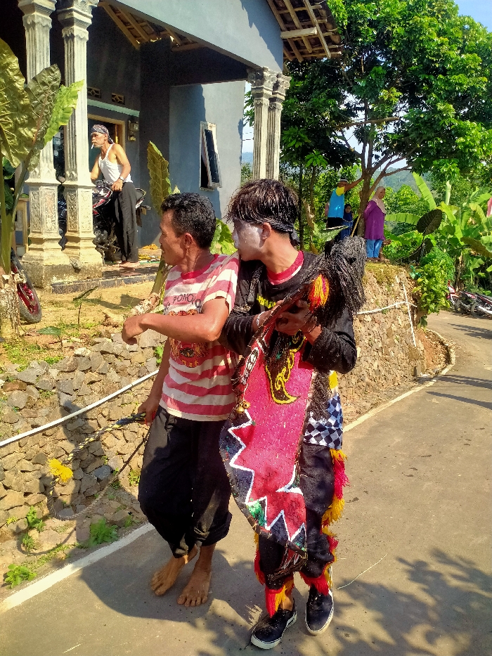 This image depicts a rural or semi-rural street scene in Jetis, Indonesia. In the foreground, two males walk along a paved road. The male on the right wears traditional Kuda Lumping attire, consisting of a black long-sleeved shirt, black pants with colorful red and yellow tassels, a black headscarf, and white face paint. He holds a decorated, horse-shaped dance prop with a dark, braided mane. He wears black sneakers with white soles and looks towards the right. To his left, another adult male, barefoot with mud on his lower legs and feet, wears a red and white striped t-shirt and dark pants. He holds a whip-like object with a yellow tuft in his right hand and looks in the same general direction.
To the left of the road, a stone retaining wall supports an elevated grassy area. A white pipe or rail runs horizontally along the top of this wall. Behind the wall, a grey-painted house with white pillars is visible. An older male with a white tank top, dark pants, and a headscarf stands by the house's entrance, leaning against a motorcycle, with a cigarette in his mouth.
In the background, on an elevated earthen mound behind the stone wall, a lush green tree provides shade. Two individuals are visible here; one female wears a purple head covering and top, and another person is partially obscured in blue and yellow attire. Hills covered in dense vegetation are visible in the distant background under a bright, sunny sky.