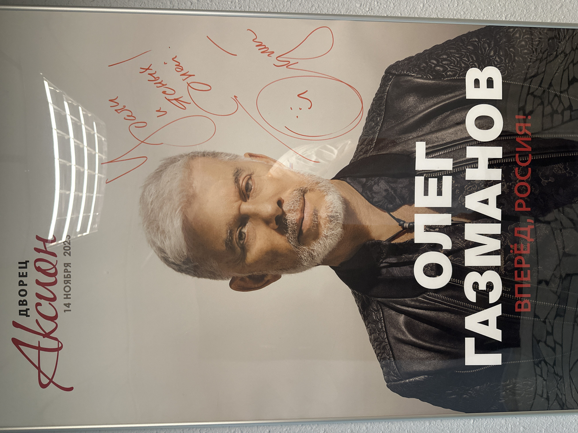 The image shows a framed poster of Oleg Gazmanov, a Russian singer, with a concert scheduled for November 14, 2023, at the Akcion Palace in Izhevsk, Russia. The poster features a portrait of Gazmanov wearing a black leather jacket. The poster also contains red signatures and notes.