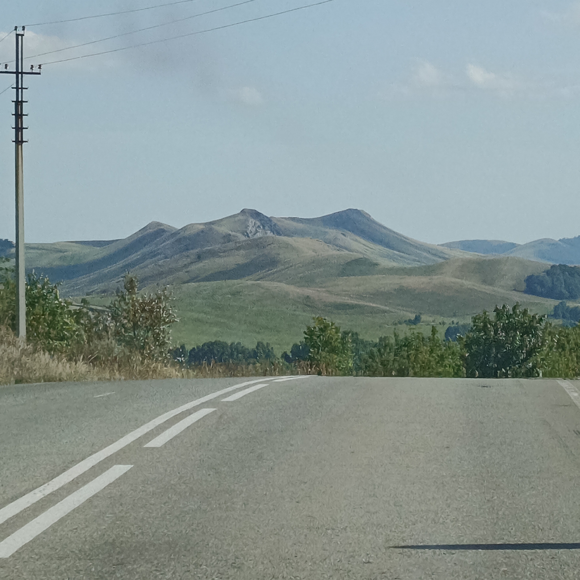 The image shows a paved road with double white lines curving towards the right. A utility pole with wires is visible on the left side of the road. In the background, rolling green hills dominate the landscape, with a distinct mountain range in the distance. The sky is clear and blue with a few scattered clouds. Vegetation, including trees and dry grass, lines the sides of the road. The scene is captured in daylight under a clear sky, likely during warmer months.